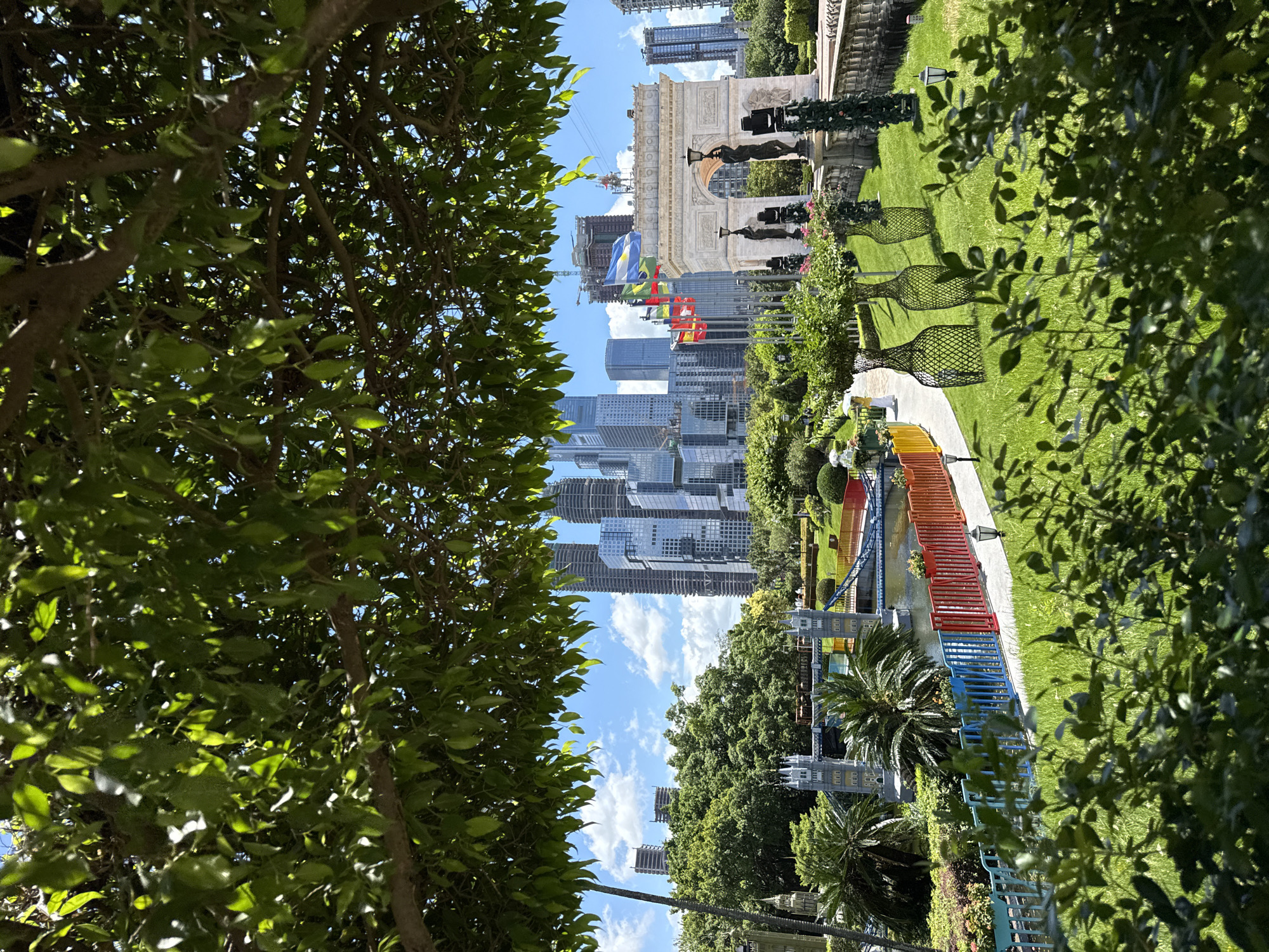 This image captures a scenic view of the Nanshan District in China, showcasing a miniature replica of the Arc de Triomphe against a backdrop of modern skyscrapers. The foreground is dominated by lush green trees and bushes, with a pathway lined by a colorful fence of blue, red, and yellow.
The miniature Arc de Triomphe is a white stone structure with statues and sculptures adorning its sides. Several flags, including those of Argentina, Uruguay, and possibly others, are planted in front of the monument, fluttering in the breeze. The overall impression is that of a meticulously landscaped park or garden.
In the background, a cluster of contemporary skyscrapers rises into the bright blue sky, dotted with fluffy white clouds. These buildings, some appearing to be under construction, represent the urban landscape of Nanshan District.
To the left of the Arc de Triomphe, a miniature replica of the Tower Bridge from London is visible, spanning a small body of water. Palm trees and other tropical vegetation add to the park-like atmosphere.
Several decorative wire sculptures resembling vases, filled with greenery and flowers, are placed on the lawn. A few lampposts are also visible, hinting at the possibility of evening illumination. A person in a yellow outfit is seen walking on the path near the colorful fence.