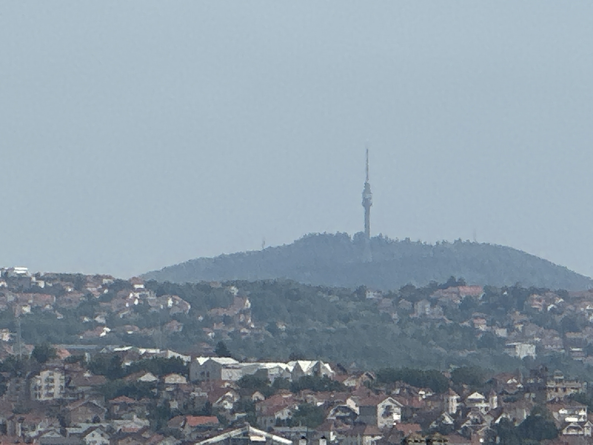 The image shows a wide shot of Belgrade, Serbia, featuring a hazy, blue sky and a densely populated hillside. The foreground is dominated by a sprawling residential area with numerous houses, many featuring red-tiled roofs. The houses are clustered together, with some larger, more modern buildings interspersed among the older structures. Green trees are visible throughout the residential area, suggesting a hilly, verdant landscape.
In the background, a large, forested hill rises, partially obscured by atmospheric haze. At the very top of this hill, a tall, slender tower is visible. This tower is likely the Avala Tower, a telecommunications tower located in Belgrade, known for its distinctive needle-like structure. The tower stands prominently against the pale sky, serving as a focal point in the distance. The overall impression is of a city built on rolling terrain, with residential development extending up the slopes towards the prominent landmark. The image appears to be taken from a vantage point overlooking the city, capturing a panoramic view.