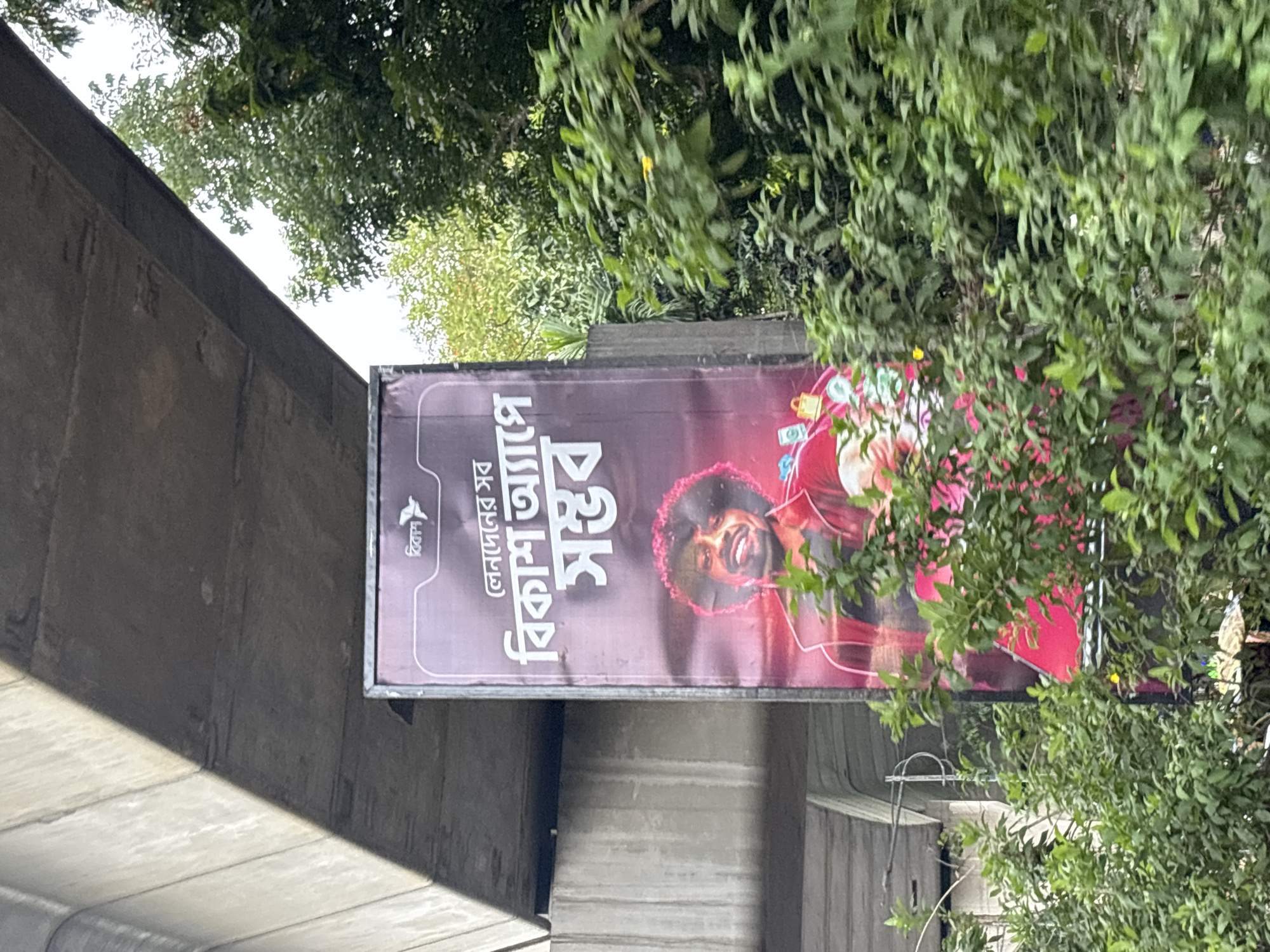 A horizontally-oriented outdoor photograph captures a large advertising billboard affixed to a concrete structure, possibly a flyover or bridge, likely in Dhaka, Bangladesh. The billboard promotes " বিকাশ " (Bkash), a mobile financial service, with the tagline "লেনদেনের সব বিকাশ অ্যাপে সম্ভব" (Lenden er sob Bikash apps-e somvob), implying that all transactions are possible through the Bkash app. A portrait of a smiling individual is prominently featured on the billboard, possibly a brand ambassador. The billboard is situated amidst lush green trees and foliage, partially obscuring the advertisement. The concrete structure to which the billboard is attached appears to be part of urban infrastructure. The overcast sky is visible in the background.