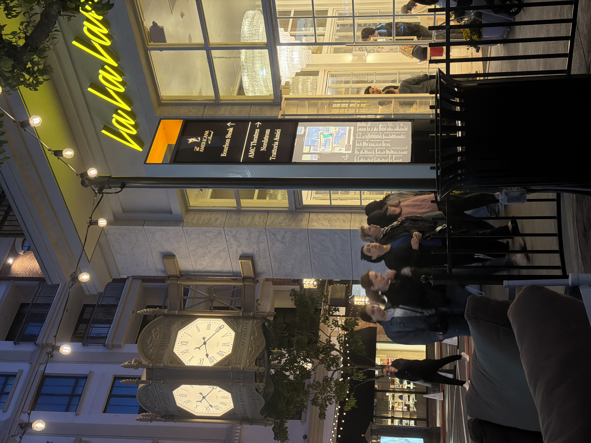 The image captures an outdoor scene at The Americana at Brand, located in Glendale, California, United States. Dominating the upper portion of the frame is a sign displaying the restaurant name "La La Land", illuminated in yellow. Below the sign, a directory indicates directions to establishments like "Bourbon Steak", "AMC Theatres", "Nordstrom", and "Trattoria Amici".
A group of people is standing on the right of the image, near a black railing. Below this, there is an outdoor lounge area. In the background, there is a clock tower in a plaza.
The environment suggests it is twilight or evening, with streetlights on. The image also captures elements of the surrounding architecture, including windows, building facades, and outdoor lighting fixtures.