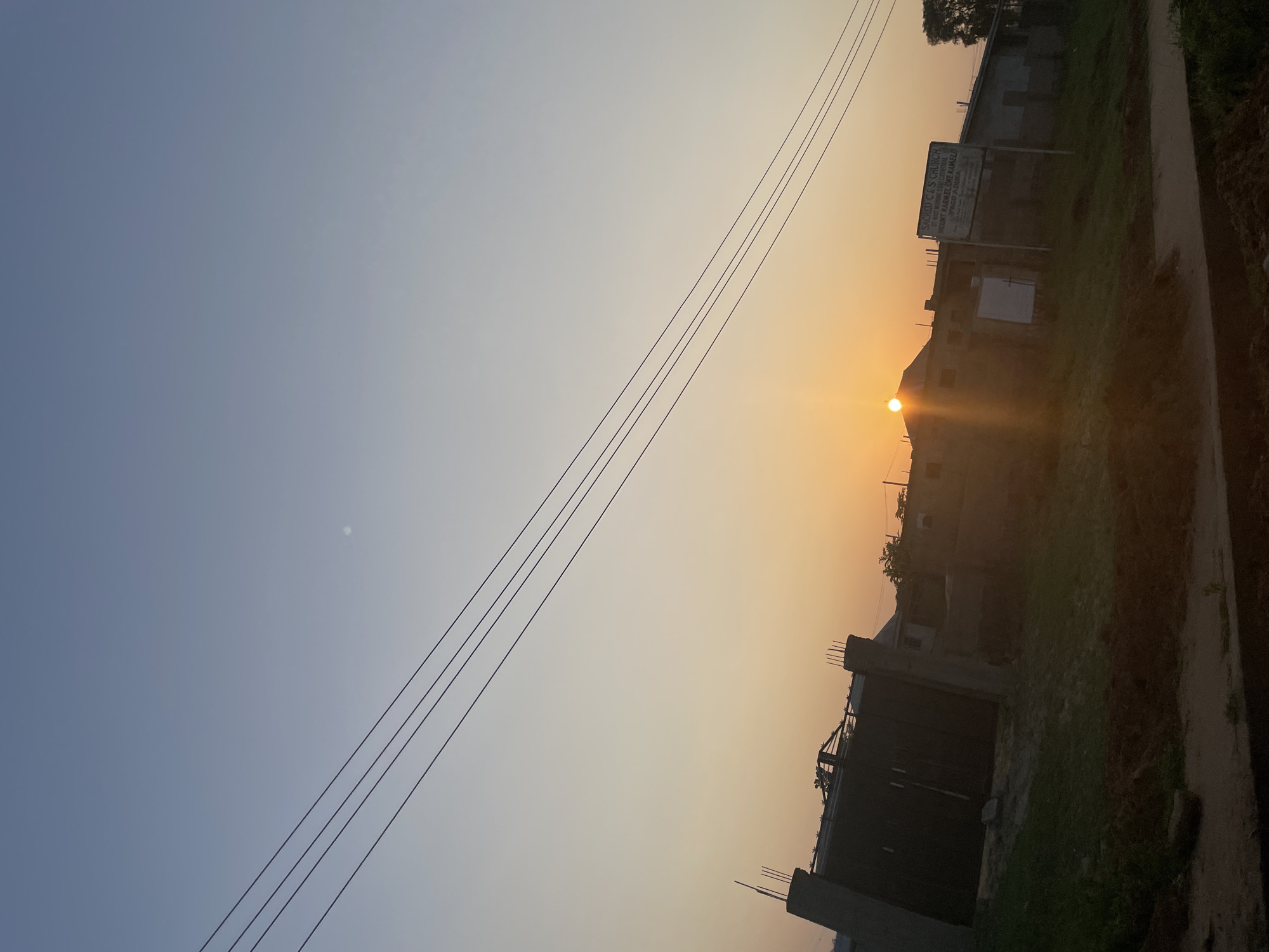 A low-angle, eye-level shot captures the warm glow of a sunrise over Iyafin, Nigeria. The sky transitions from a pale blue at the top to a soft orange and yellow gradient near the horizon. Four dark electrical wires stretch diagonally across the sky from the upper left to the lower right.
In the middle ground, several buildings are visible. To the left, a large, dark gate with a spiked fence on top dominates the scene. To the right of the gate, a longer, lower building stretches across the frame. A sign is affixed to the front of this building, though the text is not entirely legible, it appears to read "SACRED C.C.S. CHURCH." The sun, a bright orange orb, is just above the roofline of a building to the left of center, casting a bright flare and illuminating the structures with its light.
The foreground consists of unkempt, uneven ground covered in patches of grass and dark soil, suggesting a rural or undeveloped area. A dirt path or road runs horizontally across the very bottom of the frame. The overall impression is of a quiet, early morning scene in a Nigerian town. There are no people visible in the image.