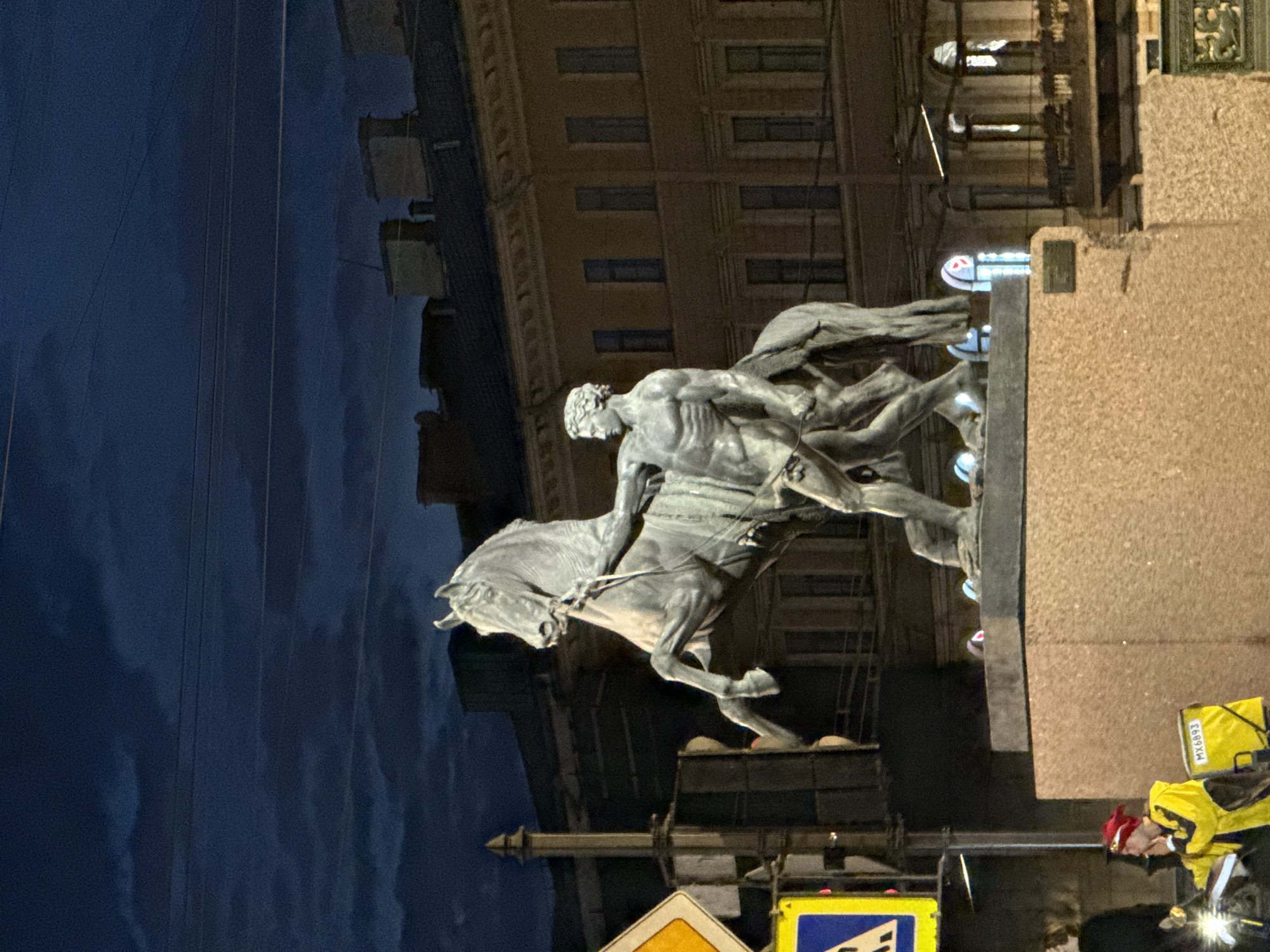 The image captures a large equestrian monument at night in an urban setting, typical of Saint Petersburg, Russia. The central subject is a majestic statue depicting a muscular male figure in a dynamic pose, riding a powerful, rearing horse. The statue appears to be made of dark metal, possibly bronze, and is dramatically illuminated by spotlights, casting clear shadows and highlighting its intricate details against the dark blue night sky.
In the background, a grand, multi-story building with numerous windows, some illuminated, suggests classical or neoclassical architecture. The sky is a deep, dark blue, indicating late evening or night, with subtle cloud formations. Thin, vertical lines, possibly overhead wires, stretch across the sky.
To the right, a large, rough-textured plinth or base for the monument fills a significant portion of the frame. In the lower right corner, a person on a bicycle or scooter, likely a delivery driver, is visible. They are wearing a bright yellow jacket and a large, matching yellow delivery bag on their back, which has the text "MX6B93" printed on it. The driver also wears a red hat and has a light on their vehicle. Partially visible street signs, one yellow and one blue, are in the lower left corner. The scene blends the historical grandeur of the monument with elements of modern city life under the artificial glow of streetlights.