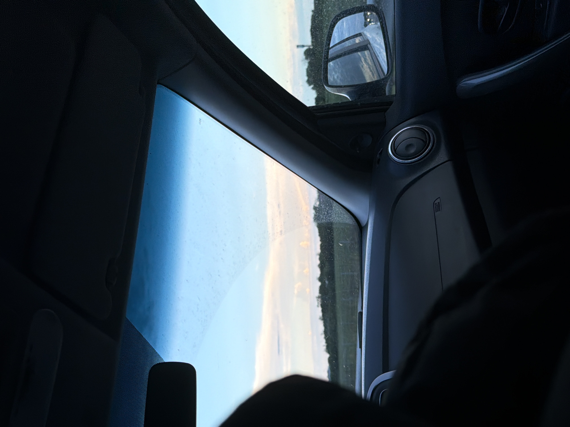 The image shows the interior of a car, looking out through the windshield. A person's dark silhouette is visible in the foreground on the left. The sky is a light blue with some wispy white and pink clouds, indicating either sunrise or sunset. A line of dark green trees forms the horizon in the distance, with a field in front of them. The car's dashboard is visible, including an air vent. A side mirror on the right side of the car shows a reflection of the road behind. The location is identified as Oktiabrskaya, Russia.