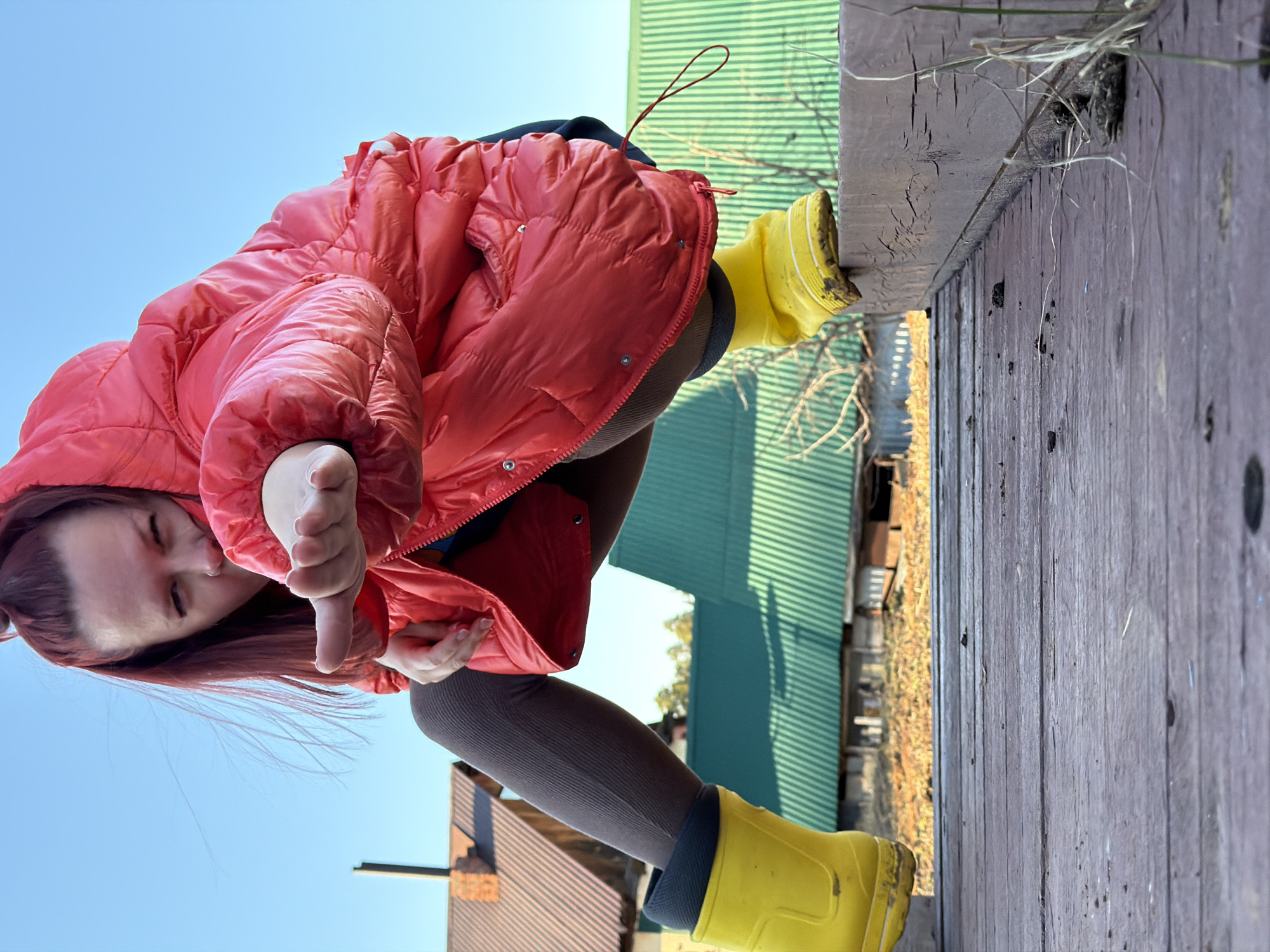 A female is shown at an extreme angle, wearing a red puffer jacket, gray leggings, and yellow rain boots, and giving a thumbs down gesture. The background includes a wooden plank structure, a green metal building, and partial views of other buildings, potentially located in Chaikovsky, Russia.