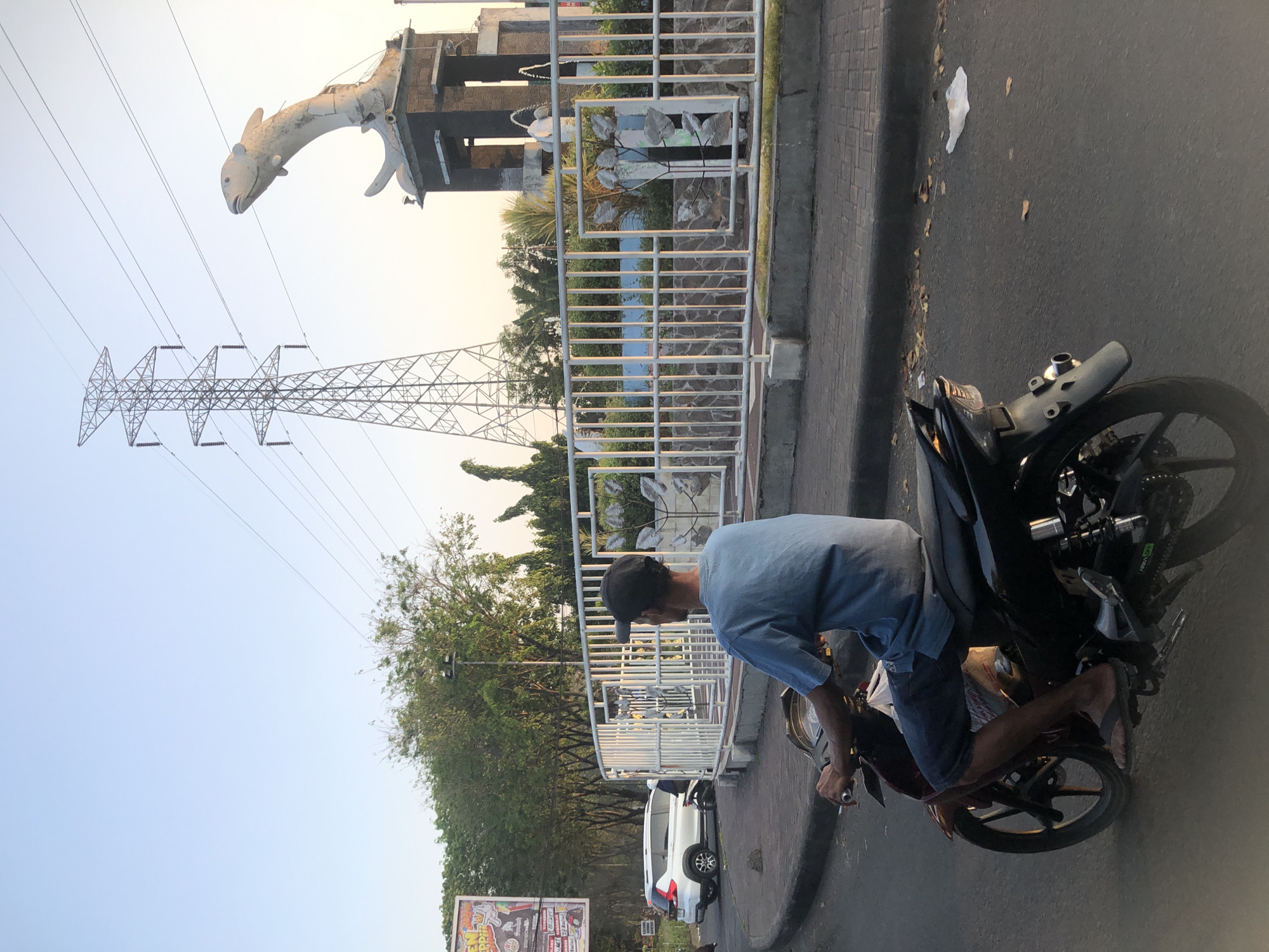 The image shows a man riding a motorcycle from behind, heading away from the camera on a paved road. He is wearing a blue t-shirt and blue shorts. The motorcycle is dark colored. In the background, there is a large metal electricity pylon and a statue of a fish. There is also a white railing with decorative elements, a white SUV parked on the left side of the road, and a billboard visible on the far left. Trees and foliage line the background. The sky is clear and light blue, indicating daytime. The scene is set in Sepande, Indonesia.