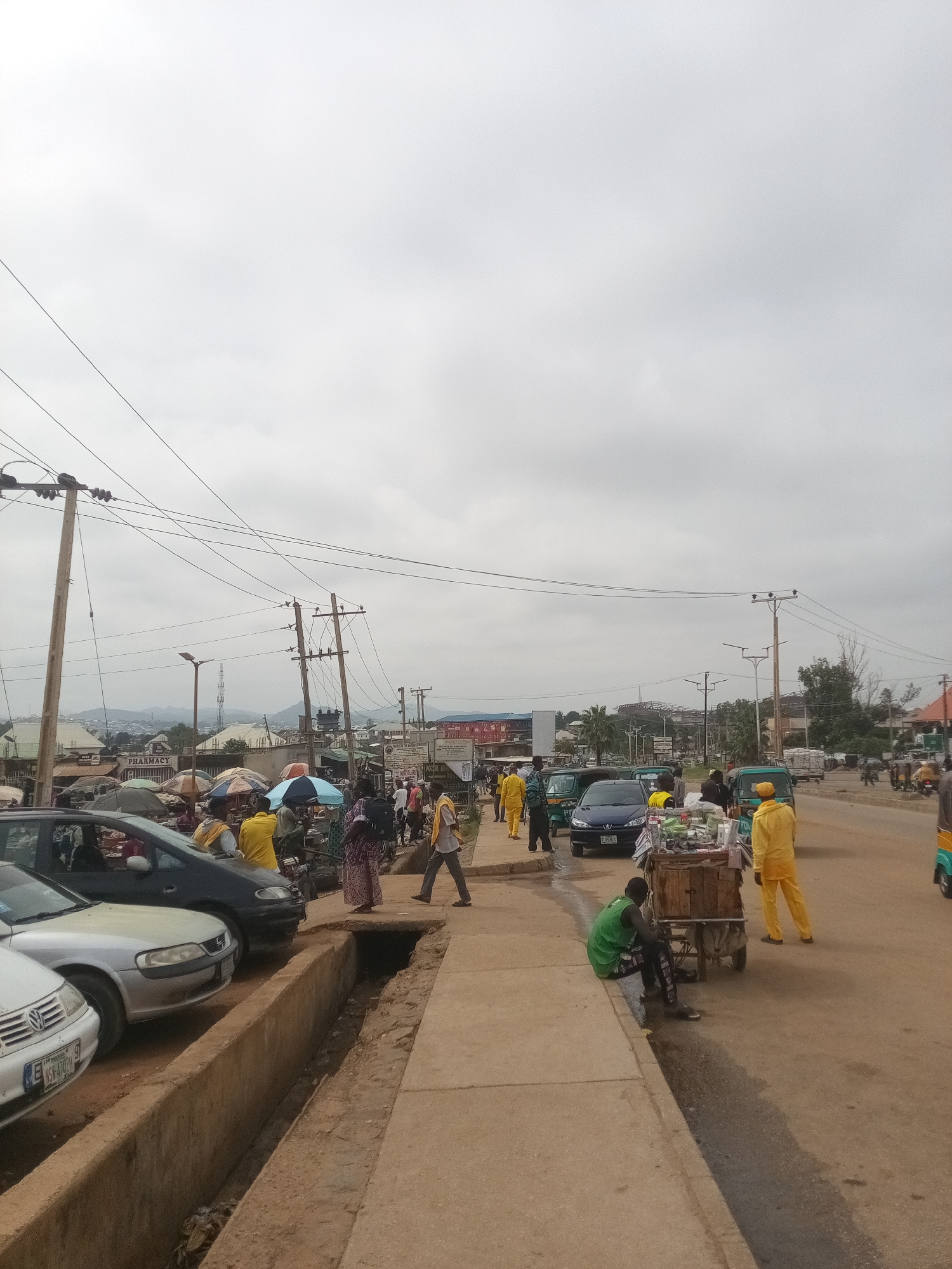 The image depicts a busy street scene in Rafin Par, Nigeria, under an overcast sky. The foreground features a concrete drainage channel running alongside a pedestrian walkway. Several vehicles, including cars and what appear to be autorickshaws, are parked or moving on the dirt road to the right. Pedestrians are walking along the walkway and interacting with street vendors. Some individuals are dressed in brightly colored clothing, including yellow. Buildings and market stalls are visible in the background, with some displaying signage. Overhead, numerous electrical wires are strung between utility poles, a common sight in urban Nigerian environments. The overall impression is one of a vibrant, active marketplace and thoroughfare.