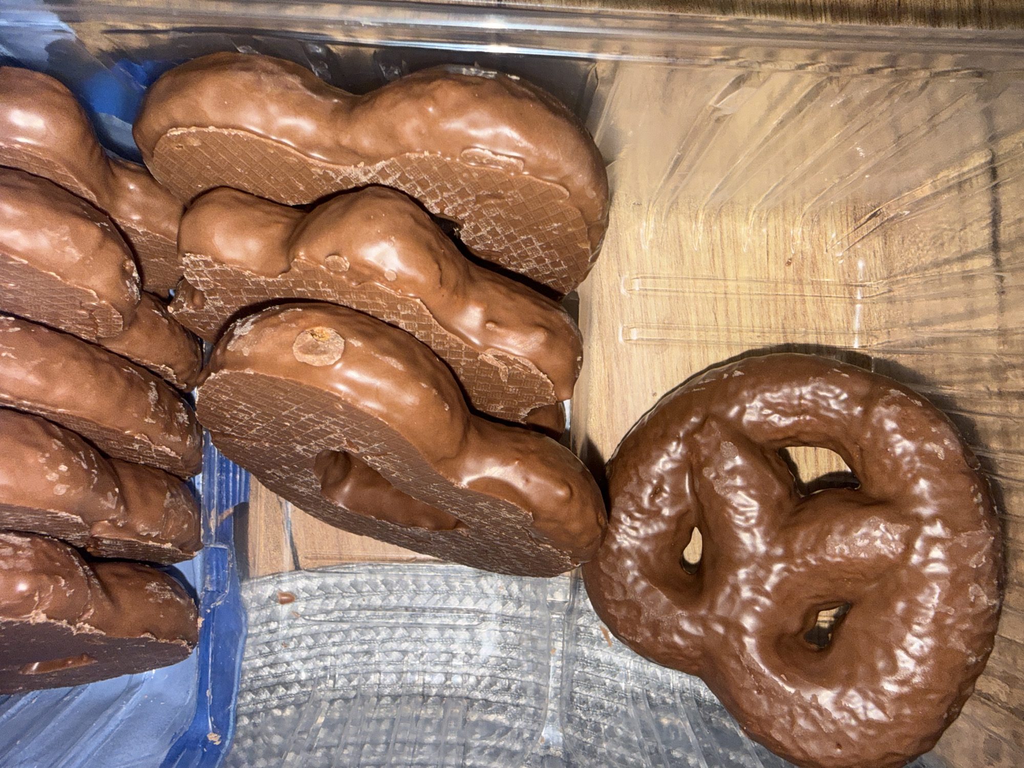 A top-down, close-up shot captures an assortment of chocolate-covered confections housed within a clear plastic container. On the left side, a stack of multiple heart-shaped or rounded treats is visible. The underside of these items clearly shows a corrugated or waffle-like texture, indicating they are wafers generously coated in a thick, rich brown chocolate. The chocolate coating on these stacked pieces appears somewhat irregular.
To the right, a single, large chocolate-covered treat shaped like a pretzel rests beside the stack. This item boasts a smoother, glossier dark brown chocolate coating, with the distinctive knot and open spaces characteristic of a pretzel.
The clear plastic container sits on a wooden surface with a visible grain. A glimpse of blue material, possibly another container or lining, is visible at the bottom left beneath the plastic box. The lighting suggests an indoor setting, likely under artificial illumination. No human presence or visible text is present in the image.
