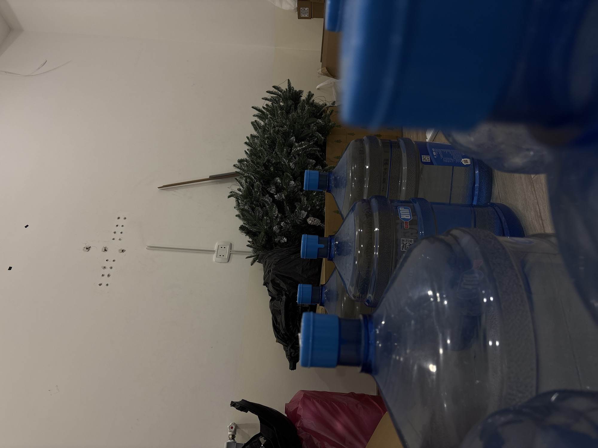 The scene is an interior view with a white wall on the left. The wall has multiple drilled holes and an electrical outlet. To the right of the wall are a few water cooler bottles, some in sharp focus and others out of focus. A small Christmas tree is standing to the left and a brown box can be seen above the tree. There are also some plastic bags, one pink and one black, on the floor. It is not possible to determine the exact location in Yerevan, Armenia, from the given visual information.
