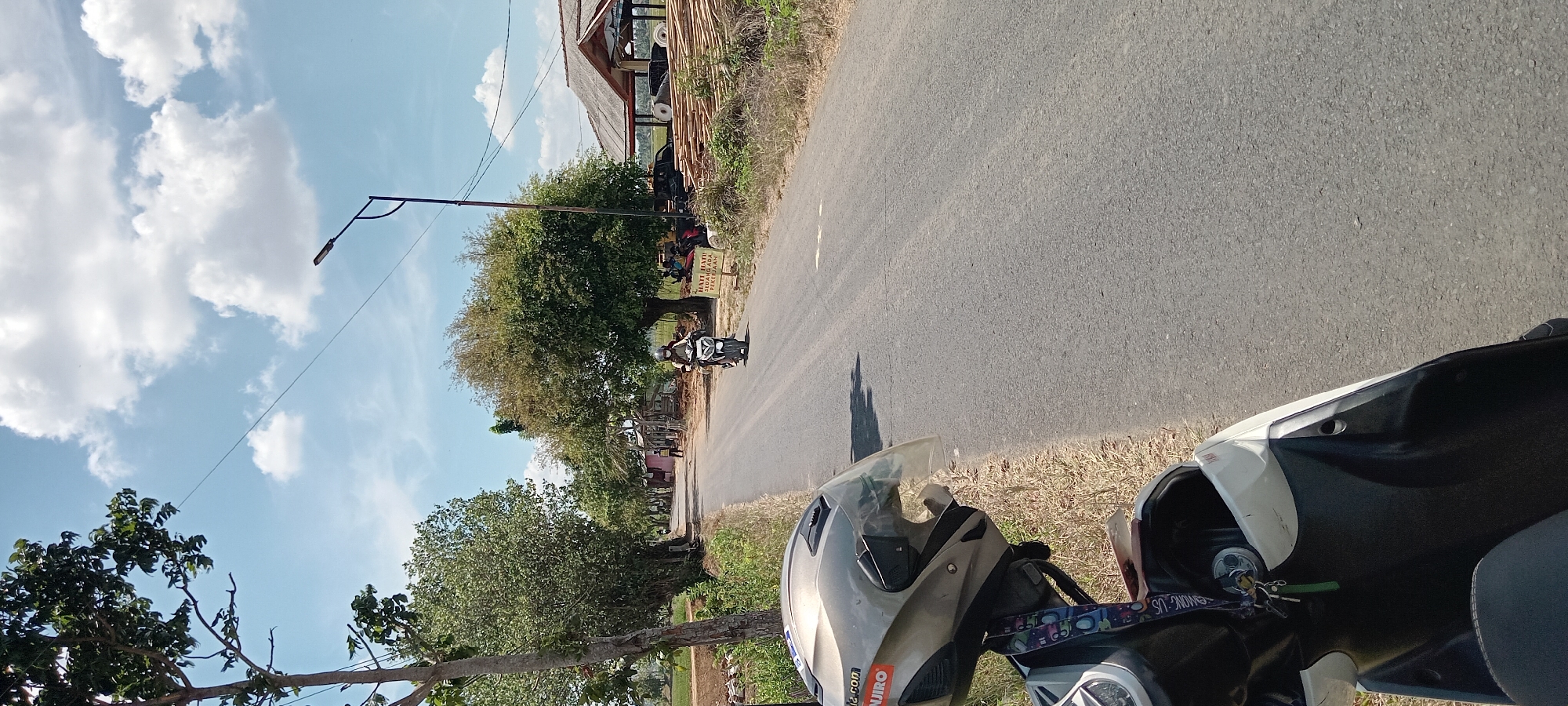 The image depicts a road scene in Paledah, Indonesia, on a sunny day with a bright blue sky dotted with white clouds. In the foreground, a white scooter is partially visible, with a silver helmet resting on its handlebars. The helmet has stickers on it, including one that appears to read "NIRO". The scooter has a lanyard with colorful cartoon characters attached to the ignition.
The road itself is asphalt and stretches into the distance, lined with trees and vegetation on both sides. On the left side of the road, there's a grassy verge and more trees. On the right side, there are several structures, including what appears to be a small open-air building with a corrugated roof, piles of lumber, and some people gathered. A sign with Indonesian text, possibly a warning or informational notice, is also visible on the right.
Further down the road, a person is riding a motorcycle away from the camera. This person is wearing a helmet and appears to be navigating the asphalt road. The overall environment suggests a rural or semi-rural setting within Paledah.