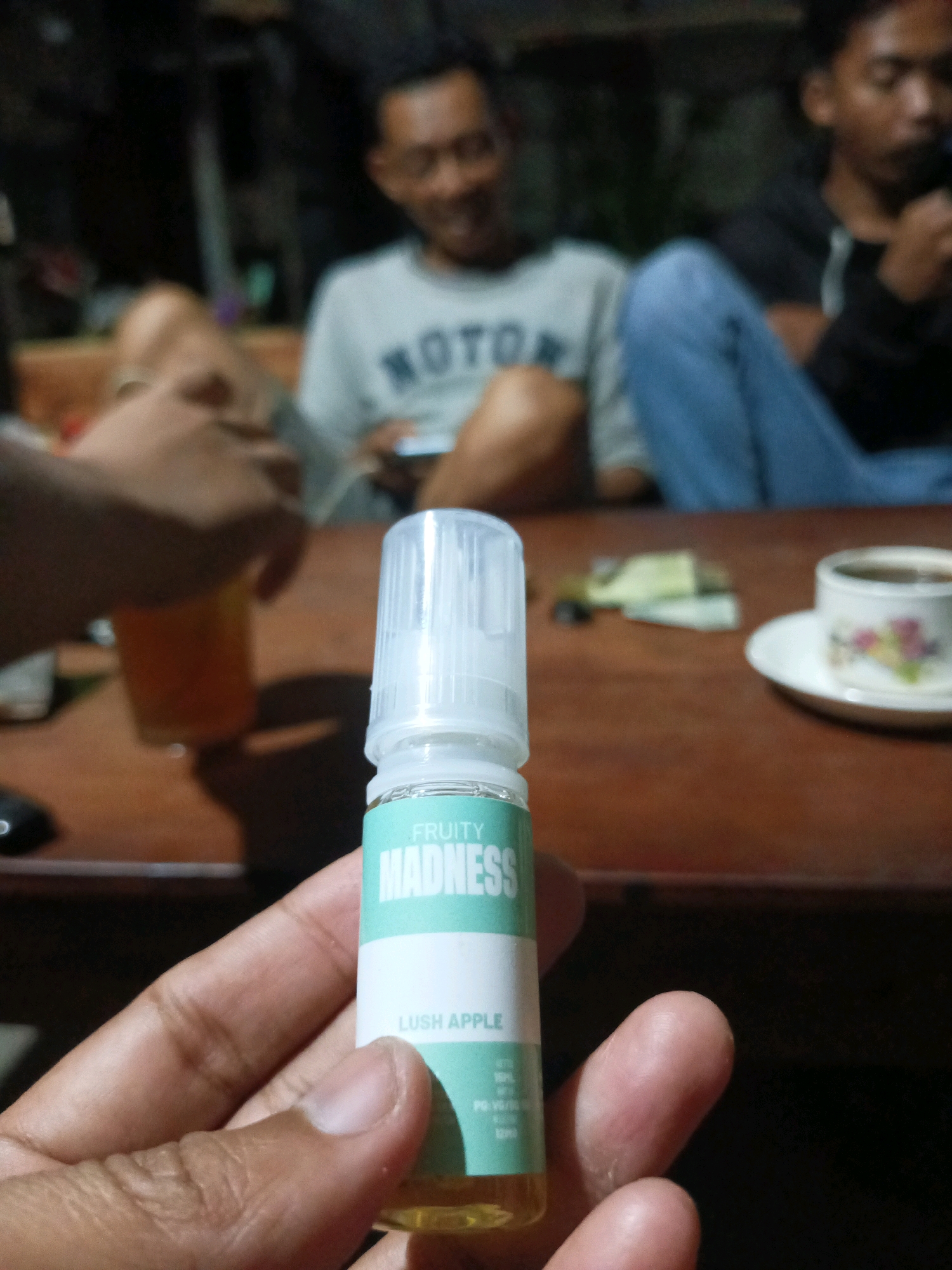 In the city of Bolorejo, Indonesia, a close-up shot focuses on a hand holding a small, clear plastic bottle with a white cap. The bottle contains a yellow liquid and features a label with green and white text. The visible text on the label includes "FRUITY MADNESS" and "LUSH APPLE". In the blurred background, three individuals are present, appearing to be seated around a wooden table. One person on the left is wearing glasses and a light-colored t-shirt with the word "MOTO" visible. Another individual in the center is wearing blue jeans. On the right, a person in a dark shirt holds something to their mouth, possibly a cigarette or vape. A cup of coffee on a saucer is also visible on the table, along with some scattered papers or money. The overall setting suggests an informal gathering in an indoor or semi-outdoor environment.