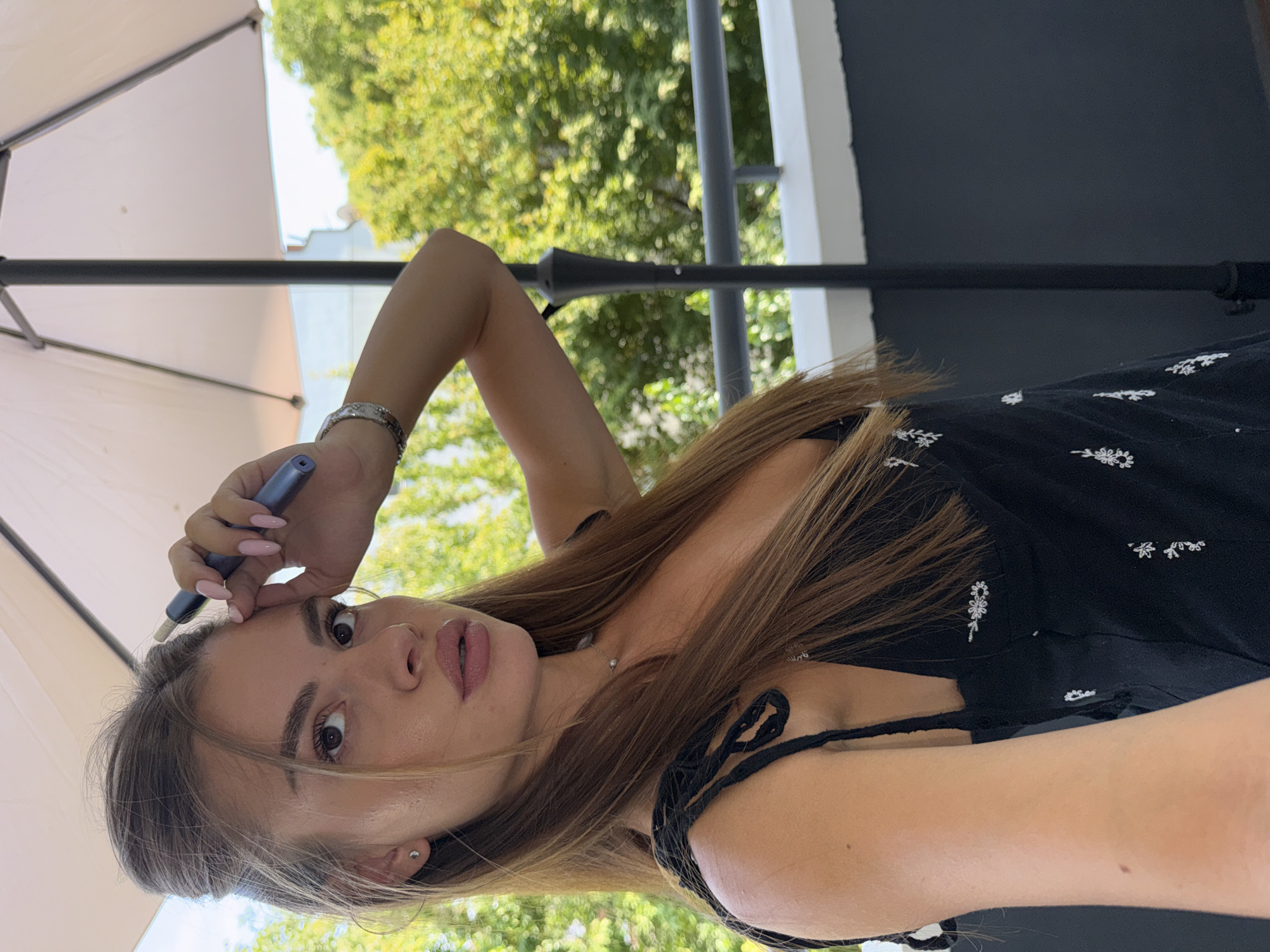 A young woman is captured in a selfie taken outdoors, possibly on a balcony, with lush green trees and a white umbrella visible in the background. She has long, light brown hair styled with a voluminous top knot and the rest of her hair cascading down. She is wearing a black dress with a delicate white floral pattern and thin straps tied at the shoulders. A thin silver chain with a small pendant rests on her décolletage. She is holding a blue electronic cigarette or vape device to her forehead with her right hand, which is adorned with a silver bracelet. Her nails are painted a light pink. The woman has prominent features, with full lips and defined eyebrows. The setting appears to be a bright, sunny day. It is impossible to determine the specific location within Plovdiv, Bulgaria from the visual information provided.