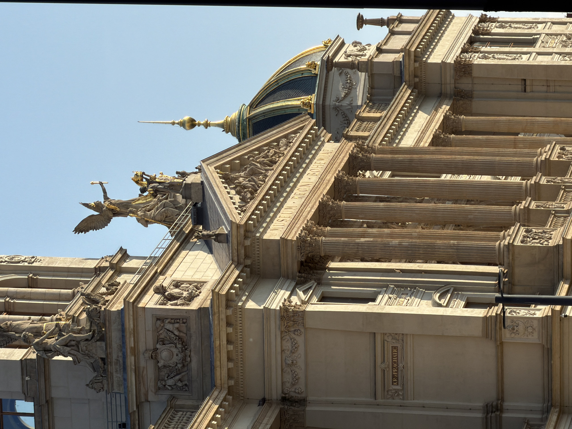 The image captures the exterior of the National Museum in Prague, Czechia, bathed in the warm light of a setting sun. The architectural style is grand and ornate, characterized by classical columns, detailed sculptures, and a prominent golden dome topped with a spire. A winged statue, likely representing victory or fame, stands prominently on the building's roof, holding a torch. The building's facade is adorned with bas-reliefs and architectural ornamentation. Inscriptions like "MUSEUM REGNI BOHEMIAE" and "KRAL>PRÁCHATICK" are visible, indicating the historical significance of the institution and potentially a section within it. The sky above is a clear, pale blue, contrasting with the warm tones of the building. The overall scene evokes a sense of historical grandeur and architectural splendor.