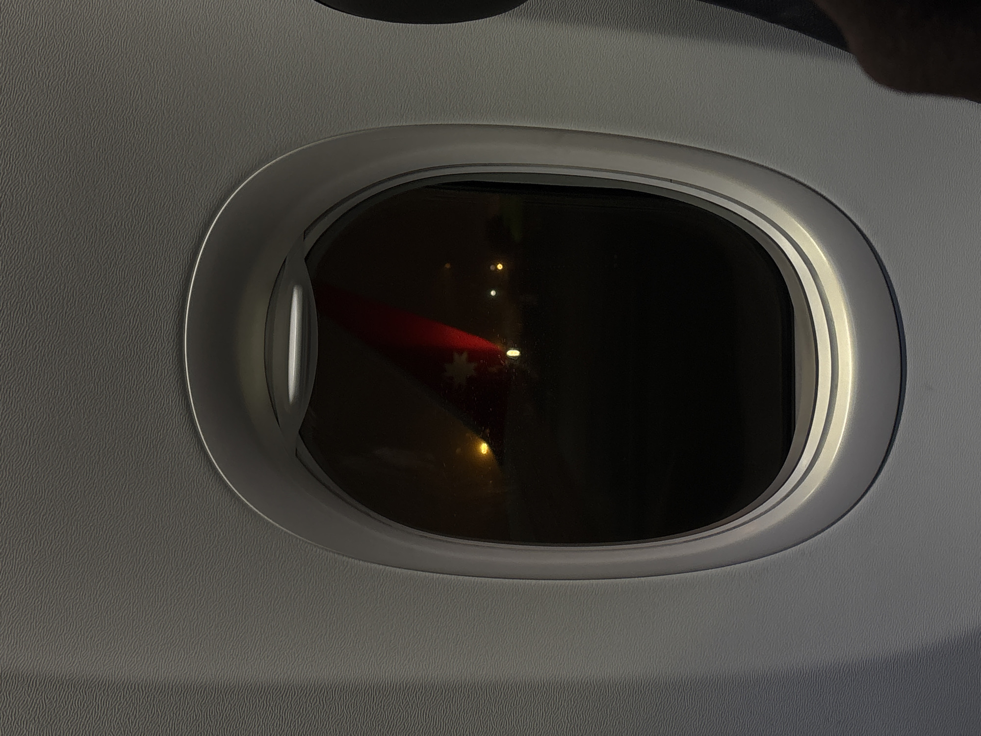 The image depicts an airplane window at night. Outside the window, there are illuminated lights on the ground and a red object, possibly part of the plane's wing or a nearby aircraft. The location could potentially be Domodedovo, Russia, assuming the reference to the city is relevant to the image's origin. There are no visible people in the frame. The setting suggests an airport environment at night.