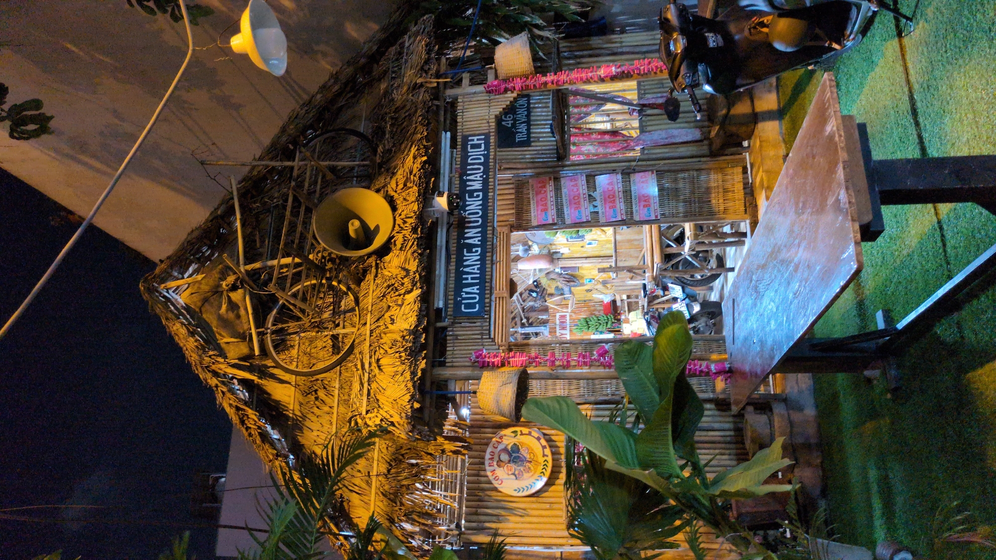 This nighttime image captures the exterior of a traditional-style establishment, identified by signage as "CỬA HÀNG ĂN UỐNG MẬU DỊCH" (General Food & Drink Store) located at 46 Trần Văn Ơn, in South Quy Nhon Ward, Vietnam. The building features a prominent thatched roof and bamboo walls. Mounted on the roof are an old bicycle and a large yellow megaphone-like speaker. A security camera is positioned above the main entrance.
To the left of the entrance, a circular sign displays "CƠM BAO CẤP" (Rationed Rice) with illustrative graphics. Woven baskets and strings of pink/red decorative elements hang near the entrance, which is framed by bamboo. Through the open entrance, the interior shows additional bamboo tables and chairs, and at least two motorbikes are parked, one partially visible inside and another black scooter parked to the right of the entrance. Menu boards featuring "CƠM BAO CẤP" are visible near the doorway.
In the foreground, a long, rustic wooden table with a dark support structure is positioned on a ground covered with green artificial turf. A large potted plant with broad green leaves occupies the left side of the foreground. A yellow pendant lamp hangs from an overhead wire, illuminating the scene. No individuals are present in the image.