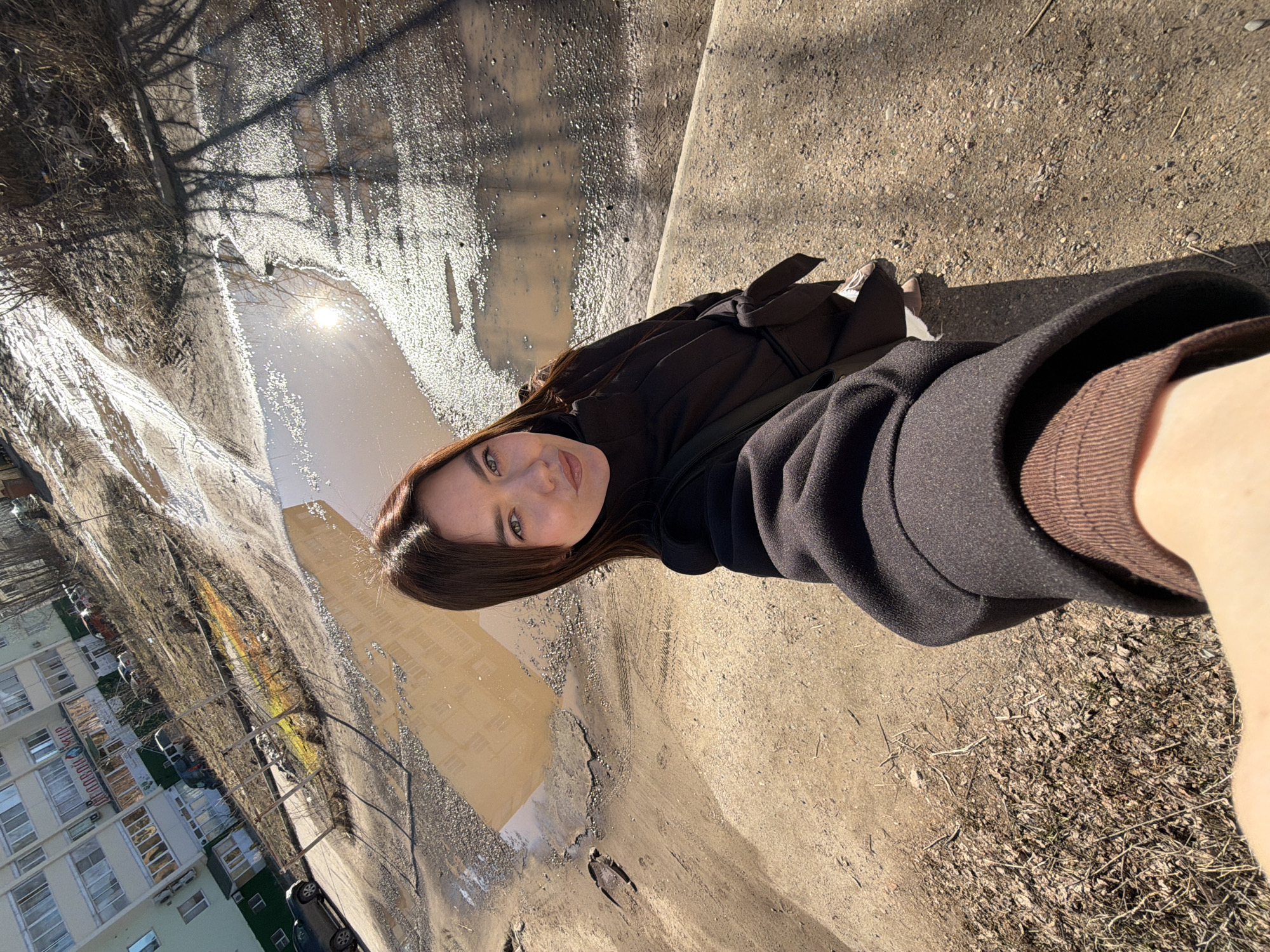 A young woman with long, dark hair and green eyes poses for a selfie on a sunny day in Chaikovsky, Russia. She wears a dark coat over a brown turtleneck sweater. The background shows a waterlogged urban street with a large puddle reflecting the sunlight and surrounding buildings. In the distance, an apartment building and parked cars can be seen. The ground is a mix of concrete and mud, indicating a post-snow melt scene.