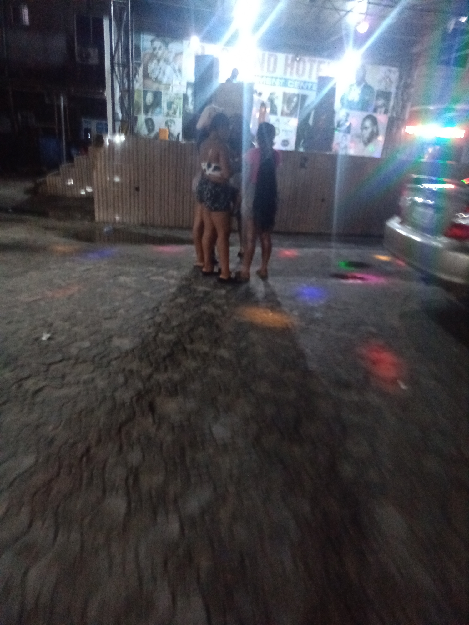 The image depicts an outdoor scene at night, likely in Ado, Nigeria, at an establishment named "ND HOTEL ENTERTAINMENT CENTRE." In the foreground, a textured, dark ground is illuminated by colorful lights scattered across it. To the right, the rear of a silver car is visible. In the mid-ground, several individuals are standing. Two women are prominently featured, facing away from the camera and towards the establishment. One is wearing a dark, patterned skirt and a white top, while the other is wearing a dark dress. Behind them, a wall with numerous framed photographs and a sign advertising "ND HOTEL ENTERTAINMENT CENTRE" is visible. The overall atmosphere suggests a lively, possibly entertainment-focused environment.