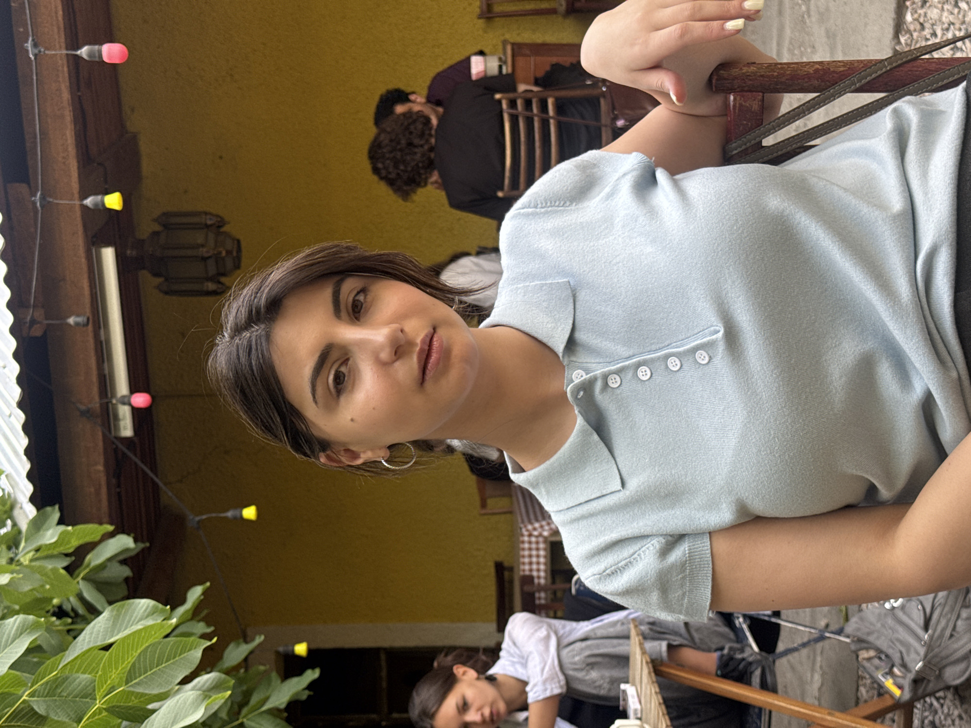 The image shows a young woman posing for a photo. She has dark hair and is wearing a light blue collared shirt with white buttons. She is sitting outdoors, with a partially visible light-yellow wall behind her. A string of lights with red and yellow bulbs is strung across the background.
In the background, other people are seated at tables. To the left, green foliage partially obscures the view. The location is likely a café or restaurant in Yerevan, Armenia.