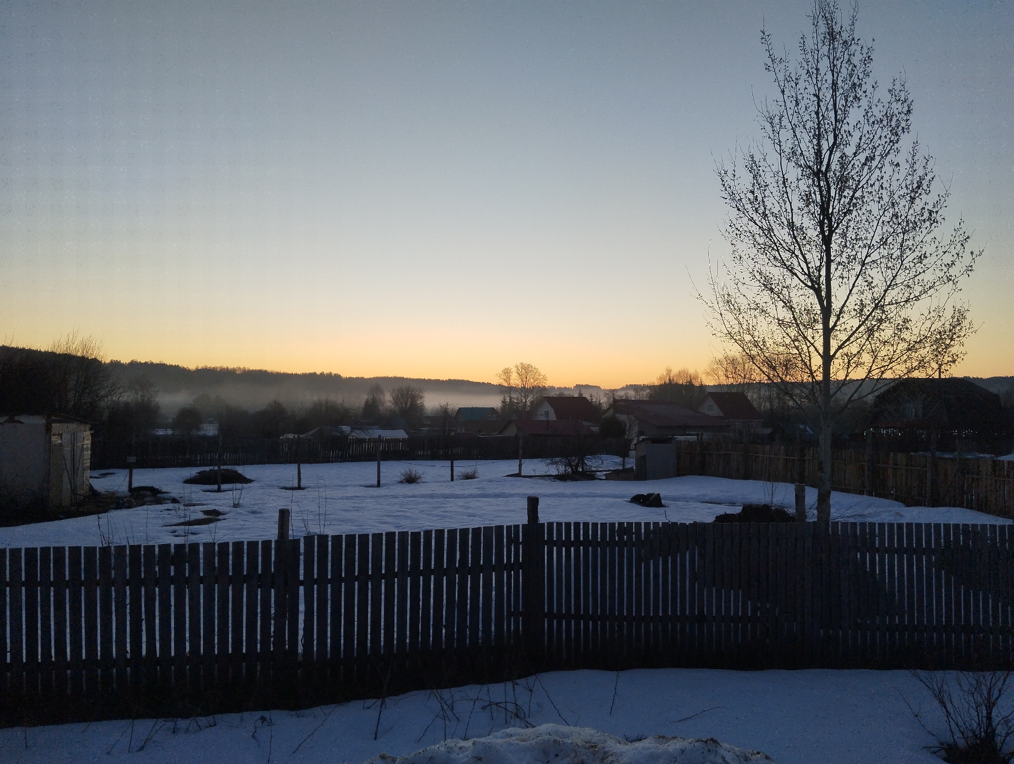 The image shows a winter scene in Grishanki, Russia at dawn or dusk. A wooden fence runs horizontally across the lower third of the frame, partially obscured by patches of snow. Behind the fence is a snow-covered field dotted with small plants and some debris. In the background are several houses and structures, silhouetted against a light orange and yellow horizon. A layer of mist or fog is present near the horizon line. A bare tree stands prominently on the right side of the image. The sky transitions from orange to a lighter shade of blue or gray toward the top of the image. The lighting suggests either sunrise or sunset. There are no visible people.