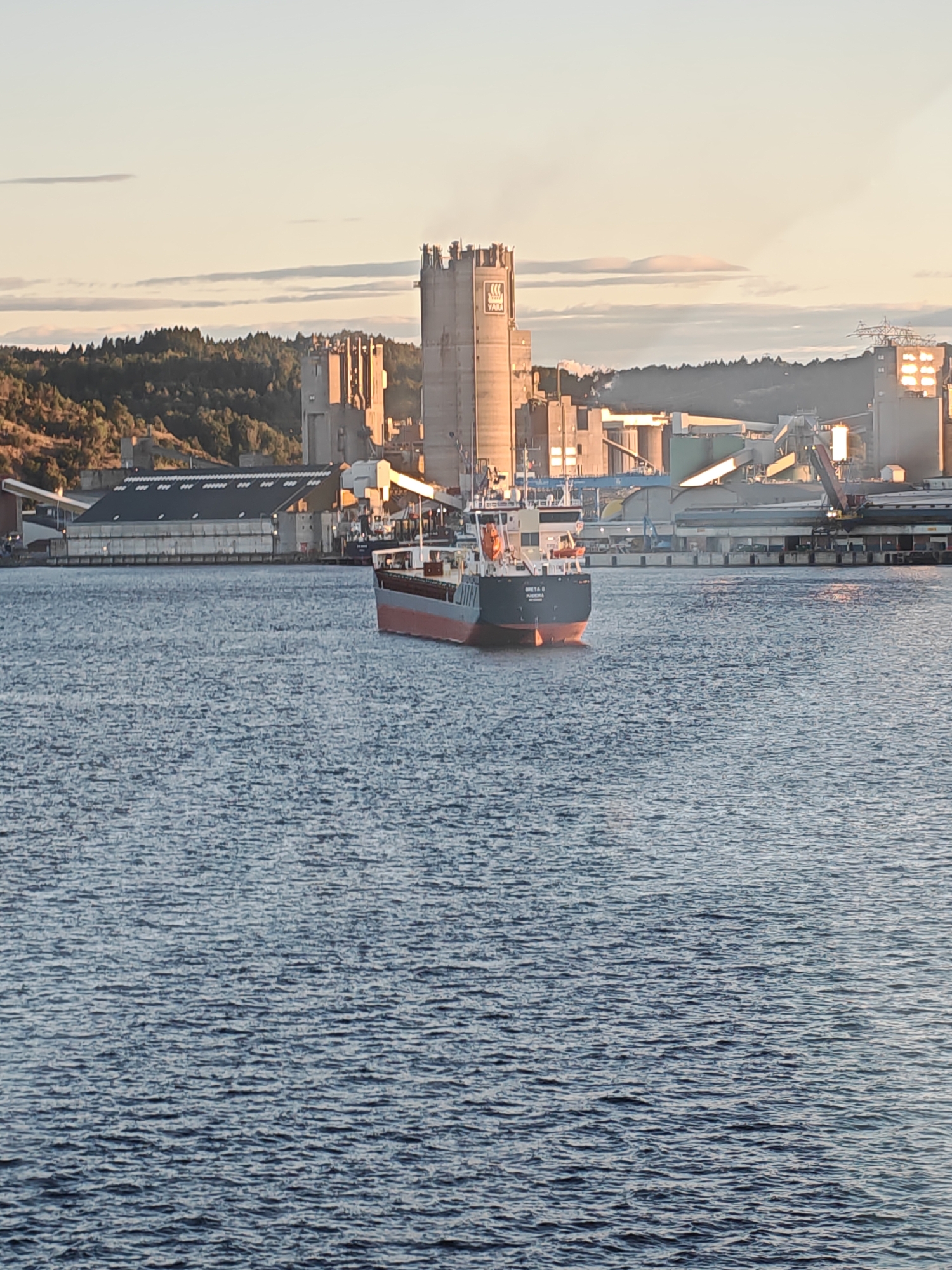 This image shows a ship sailing in the water, with an industrial area on the shore. The ship is a medium-sized cargo vessel with a red and blue hull and a white superstructure. It is sailing away from the camera, towards the industrial area. The industrial area consists of several large buildings, including a tall, cylindrical silo with the Yara logo on it. There are also cranes and other industrial equipment visible. The scene is set against a backdrop of a forested hill and a sky with some clouds. This industrial area is located in Porsgrunn, Norway, specifically at the Herøya industrial park.