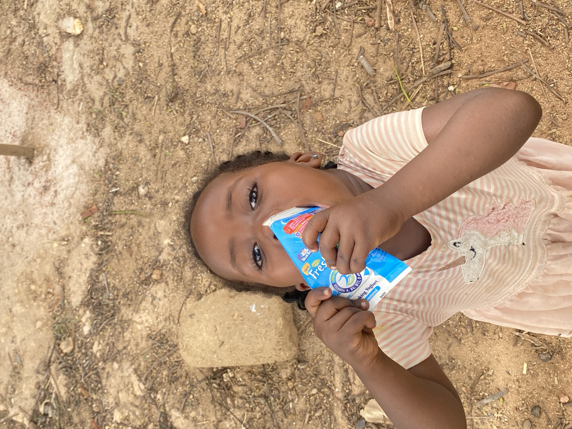 A young girl is lying on the ground in what appears to be the city of Kuru Dungus, Nigeria. She is wearing a light-colored, striped dress with a pink unicorn design on the front. She is drinking from a blue and white container of "Fresh" drinking yogurt. The ground is dirt with scattered twigs and a small rock near her head.