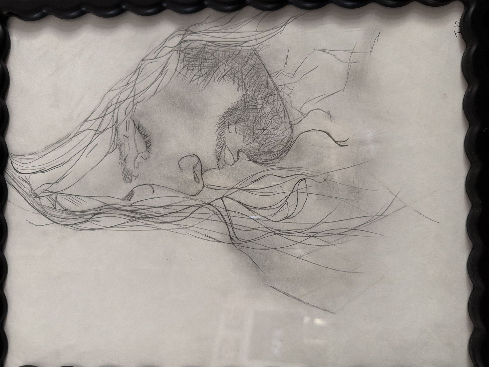 This is a framed pencil sketch of a man's head and shoulders, facing left in profile. The man has long, flowing hair, closed eyes, and a beard. The sketch is done on a light-colored paper and appears to be unsigned or signed with initials in the bottom right corner. The frame is black with a scalloped edge. The image is taken indoors, as indicated by the reflection of what appears to be indoor lighting and possibly furniture on the glass covering the artwork. There is no discernible connection to the city of Wellford, United States of America, in the image itself.