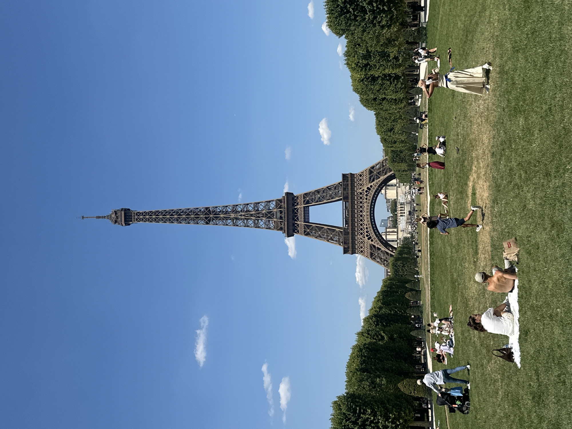 The image captures a bright, sunny day at the Champ de Mars in Paris, France, with the iconic Eiffel Tower dominating the background. A clear blue sky with a few scattered white clouds serves as the backdrop. In the foreground, a lush green lawn is dotted with people enjoying the pleasant weather.
Several individuals and groups are scattered across the lawn. Some are sitting on blankets, picnicking, while others are standing or walking. A person with a stroller is visible on the left, and a child runs across the grass in the middle of the frame. Further back, more people are seen lounging and socializing. The Eiffel Tower, made of intricate iron latticework, rises majestically in the distance, centered within a tree-lined avenue that leads towards it. The trees are full and green, suggesting a warm season. In the far background, a few buildings are visible, indicating the urban setting of Paris. The overall atmosphere is one of leisure and relaxation.