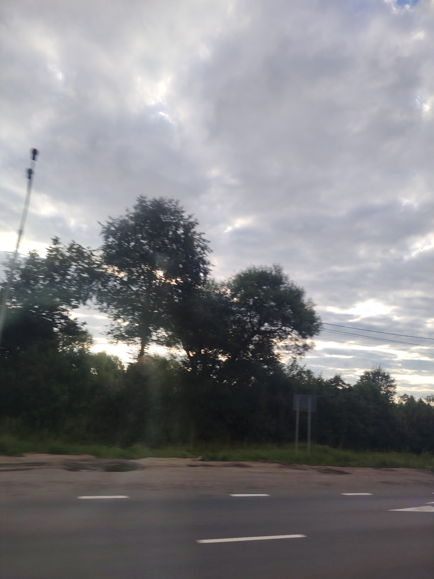 The image depicts a cloudy sky over a tree-lined road in Spas-Zaulok, Russia. The foreground shows a section of an asphalt road with white lane markings. To the right of the road, there is a dirt shoulder and a dense line of dark green trees. In the middle ground, silhouetted trees with visible sunlight filtering through their branches are prominent. A traffic sign on two grey poles is visible in the background to the right of the trees. In the far upper left, a blurred, tall, dark object, possibly a street light or utility pole, is present. The overall lighting suggests it is either early morning or late afternoon.