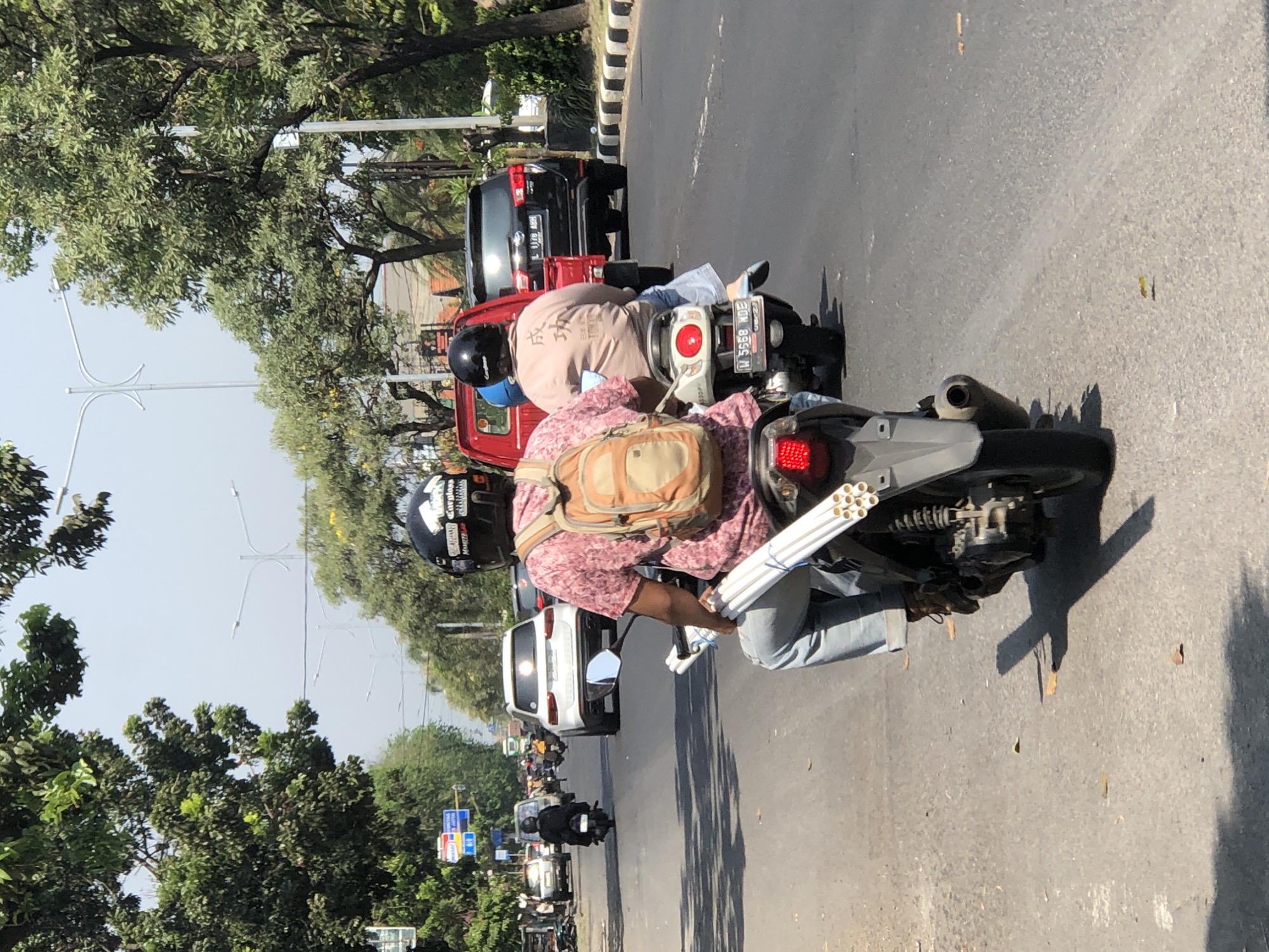 In a street scene in Larangan, Indonesia, a person is riding a motorcycle carrying a bundle of white pipes strapped to the rear of the vehicle. The rider, wearing a pink patterned shirt and jeans, has a backpack on and a black helmet. They are in the middle lane of a paved road with other vehicles visible. To the left, a white SUV is stopped at an intersection, and further down the road, several other cars and motorcycles are in motion. Trees line both sides of the street, and overhead wires are present. The sky is clear and bright. A second motorcycle carrying two people is to the right of the primary subject, also moving forward.