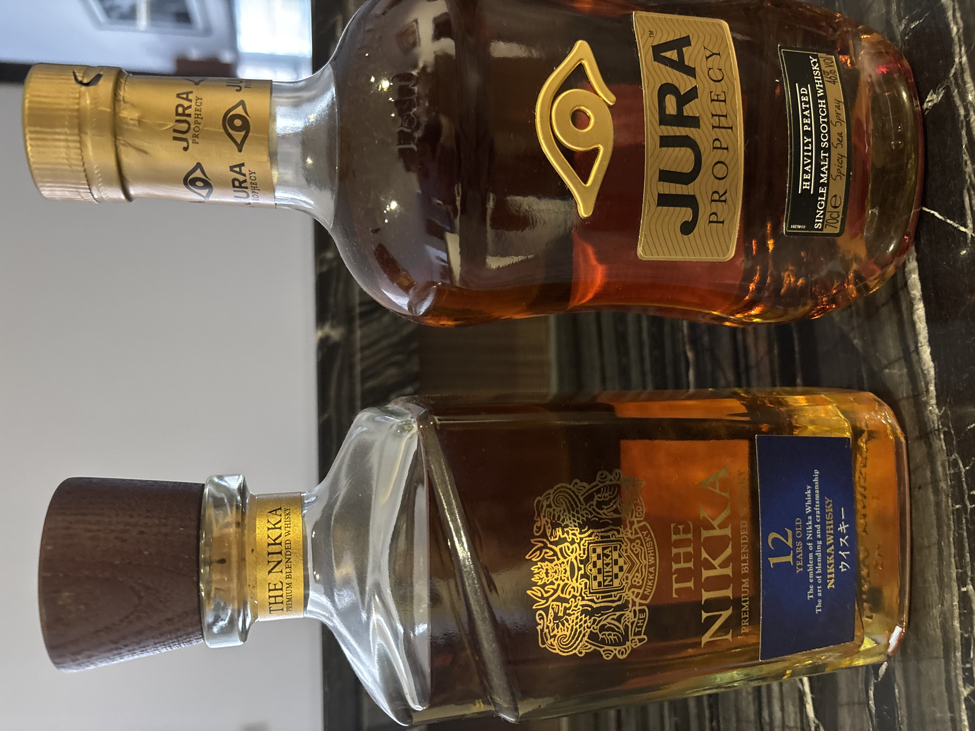 Two bottles of whiskey stand on a dark, marbled surface. The bottle on the left is The Nikka Premium Blended 12 Years Old, featuring a wooden cap and a gold crest with the Nikka logo. The bottle on the right is Jura Prophecy Single Malt Scotch Whisky, identifiable by its gold foil cap and a prominent golden eye emblem on the label. The background is blurred, showing hints of a wall and possibly artwork. There are no people or animals present in the image. The lighting suggests an indoor setting, likely a bar or a home display. The image was taken in Dubai, United Arab Emirates.