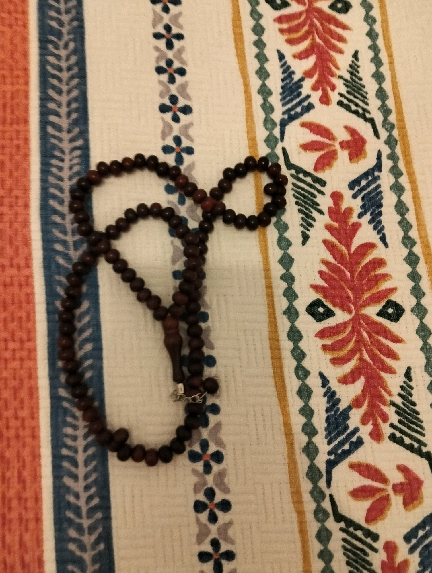 A string of dark, round beads is draped on a striped fabric. The fabric has vertical stripes of orange, white with blue floral patterns, and a cream color with a red and blue floral pattern.