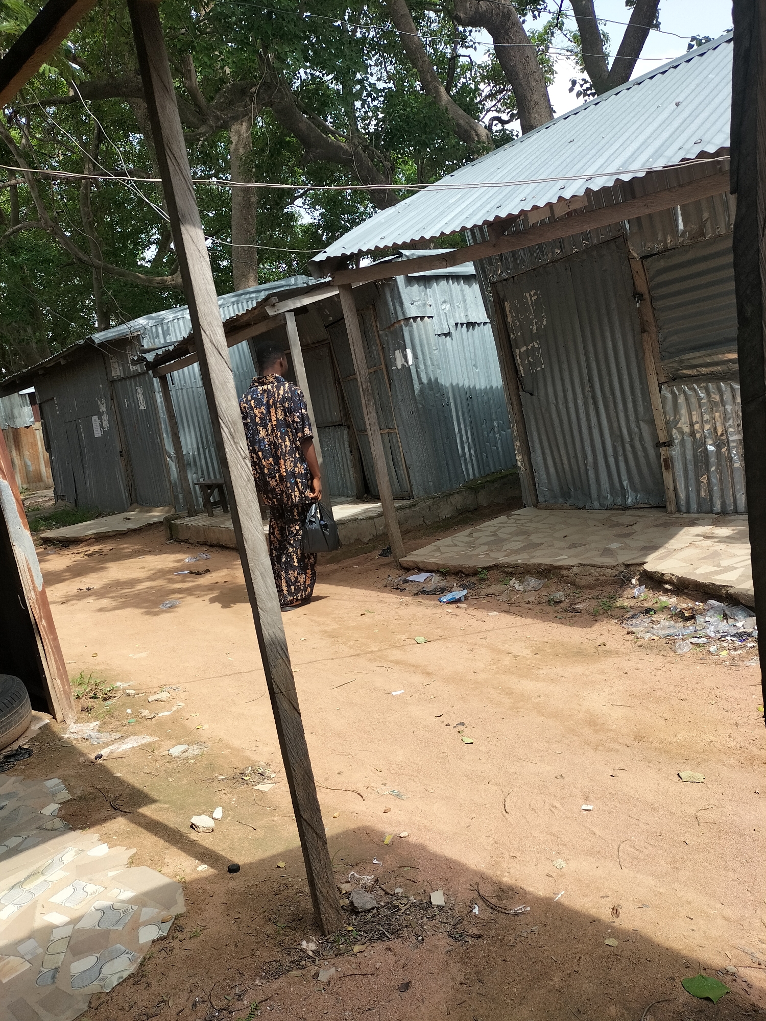 The image shows a person in a patterned garment walking away from the camera in an outdoor setting that appears to be a market or a residential area with corrugated metal structures. The ground is sandy and dusty, with some debris and scattered pieces of plastic. There are also some paved areas with tiles. In the background, there are trees with dense foliage and a corrugated metal roof on one of the structures. The scene is lit by daylight, and shadows are cast on the ground. The overall impression is of a humble, possibly informal, commercial or living space. This scene is located in Jalingo, Nigeria.