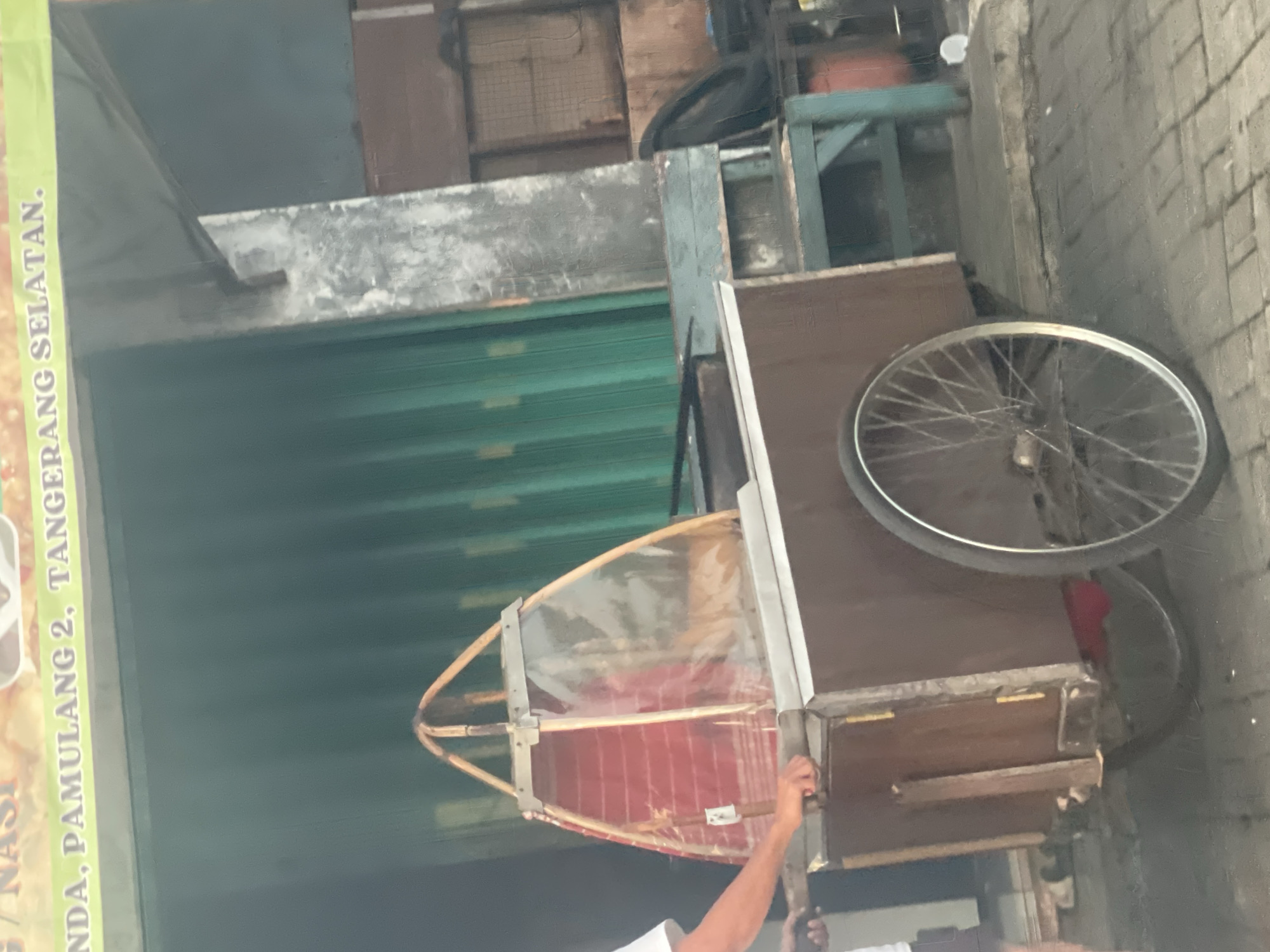 A street vendor's cart is parked on a paved surface in front of a building with a green metal shuttered facade. The cart is made of wood and has a large spoked wheel on the right side. The front of the cart is covered with a clear plastic sheet, revealing a red and white striped fabric underneath. A person's arm, wearing a white shirt, is visible on the left side of the image, reaching towards the cart. Above the cart, a banner with text is partially visible, with some words readable including "NDA, PAMULANG 2, TANGERANG SELATAN." This indicates the location is Pamulang 2, South Tangerang, Indonesia. The environment appears to be an outdoor commercial area, likely a street or sidewalk.