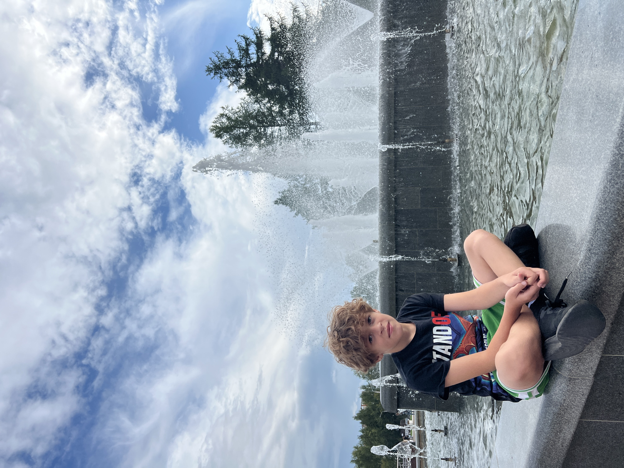 A young boy with curly light brown hair sits cross-legged on the edge of a fountain in Chaikovsky, Russia. He wears a black t-shirt with a graphic design and green shorts with white stripes. His hands are clasped in his lap, and his gaze is directed towards the camera. Behind him, a large fountain sprays water into the air, with trees and a cloudy sky visible in the background. The fountain's basin is filled with rippling water.