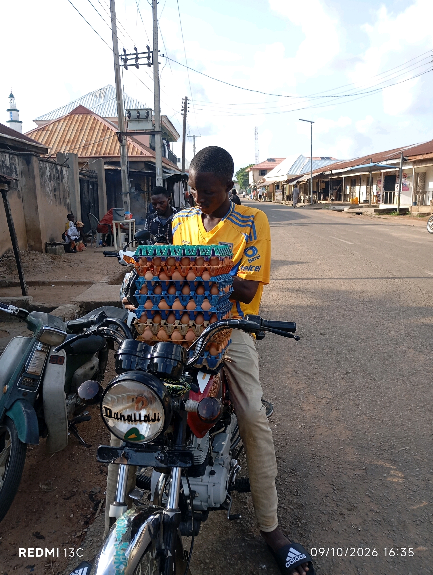 In the city of Shendam, Nigeria, a young boy, dressed in a yellow and blue T-shirt and beige pants, sits astride a motorcycle. The motorcycle is loaded with stacked trays of brown eggs. Another motorcycle, a greener model, is parked to the left. In the background, a row of shops lines a dusty road. A mosque with a tall minaret is visible in the distance. The sky is cloudy. The timestamp indicates the photo was taken on October 9, 2026, at 16:35.