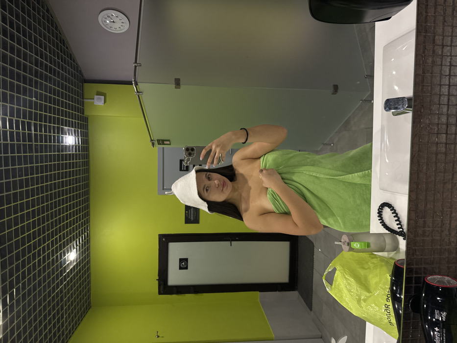 This image depicts a female subject taking a mirror selfie in an interior space, likely a changing room or spa area. The specific location in Vniissok, Russia, cannot be determined from the visual information provided.
The subject is a young adult with long, dark hair, wearing a white, conical felt sauna hat and wrapped in a bright lime-green towel covering her torso. She holds a silver-colored smartphone in her right hand, angled to capture her reflection, while her left hand rests on the towel across her chest. A black elastic band is visible on her left wrist. Her expression is neutral as she looks towards the phone's camera.
The background features a room with walls painted in contrasting colors: a vibrant lime-green on the left and a light purple-grey on the right, beyond a frosted glass partition. The ceiling is a dark, square grid with recessed lighting. To the far left, a dark-framed white door with a handicap accessibility symbol is visible. To the right, frosted glass partitions indicate shower or changing stalls. A white analog wall clock on the purple-grey wall displays approximately 1:50.
In the immediate foreground, a white countertop with two sinks and a chrome faucet is present. Resting on the counter are a partially visible bright lime-green plastic bag, a clear water bottle with a matching lime-green cap, a black coiled elastic, and part of a black hairdryer. The floor consists of light grey tiles, with a black mat visible in front of the door on the left.