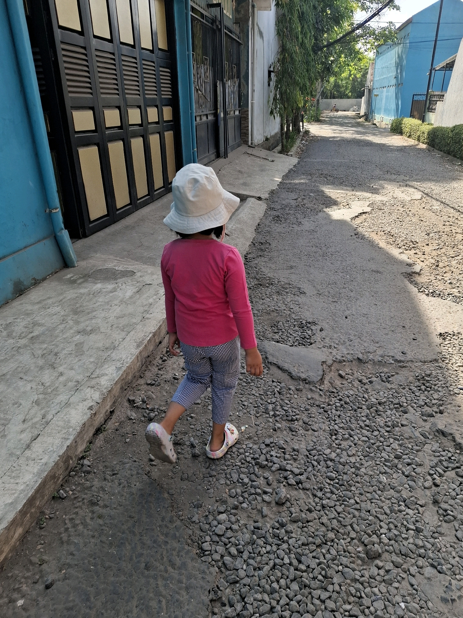 A young girl, viewed from behind, walks away from the camera down a paved alley in Karangtengah, Indonesia. She wears a pink long-sleeved shirt, striped pants, and a white bucket hat. The alley is lined with buildings and a gate on the left, and houses on the right. The ground is covered with gravel and patches of cracked asphalt. Sunlight casts shadows across the alley.