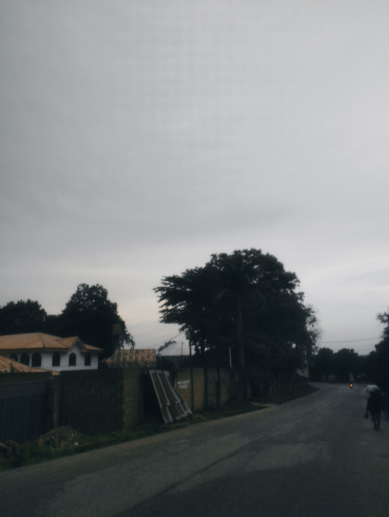The image depicts a street scene in Jos, Nigeria, under a heavily overcast sky. On the left side of the street, a white building with a light brown, sloped roof and arched windows is visible, partially obscured by a dark, textured wall. Adjacent to the building, a large, dark, leafy tree dominates the mid-ground. A paved road runs from the foreground to the background, curving to the right. A lone individual, seen from behind, walks away from the viewer on the right side of the road, carrying a white object over their shoulder. They are silhouetted against the dull light. A distant, faint orange light is visible on the right side of the road, possibly from a vehicle or street lamp. The overall scene is rendered in muted, desaturated tones, suggesting either early morning, late evening, or a day with poor lighting conditions.