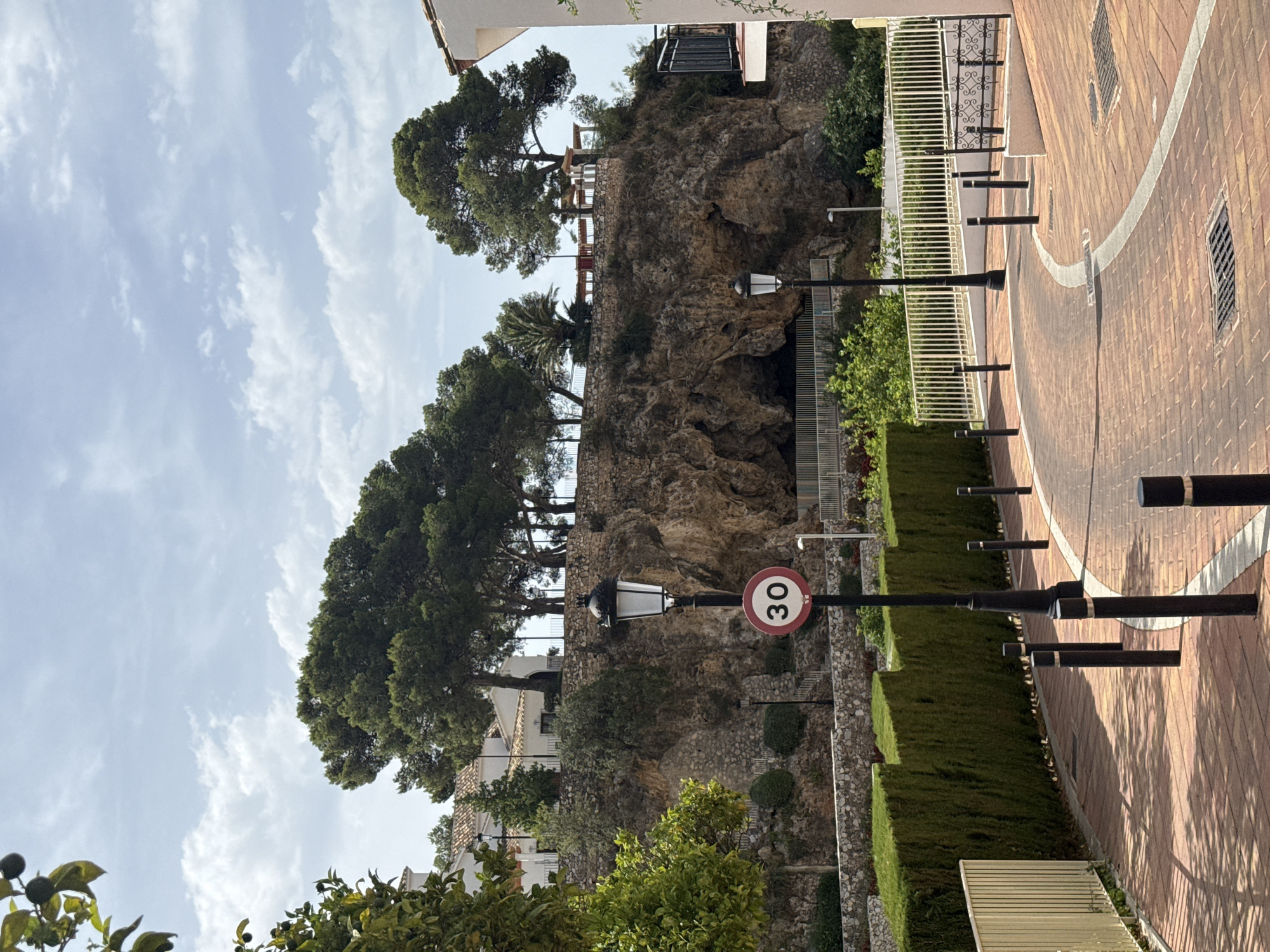 The image captures a scenic view of a street in Benalmádena, Spain, with a rocky cliff face prominently featured in the background. A winding brick-paved road, bordered by black bollards and a white line, curves through the foreground. A neatly trimmed green hedge runs along the left side of the road, separated by a low stone wall.
At the top of the cliff, several large pine trees and some palm trees are visible, along with white buildings with terracotta roofs. A dark opening, possibly a cave or tunnel, is present in the rock face. A street lamp with a decorative top stands near the road, accompanied by a circular speed limit sign displaying "30" within a red border. Another street lamp is visible further up the cliff. The sky is partly cloudy, with a mix of white clouds and blue sky. The scene appears to be bathed in daylight, with shadows indicating the sun's position. No people are visible in the image.