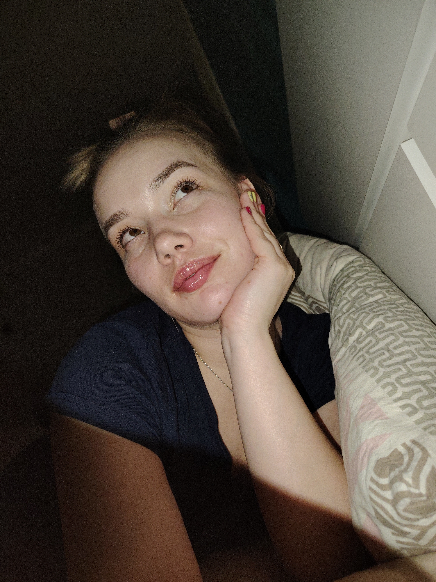 A female subject is depicted in an indoor, dimly lit environment, within the city of Марковский, Russia. She is shown from a low-angle perspective, partially reclined, wearing a dark blue short-sleeved top and a thin, silver-colored chain necklace. Her head is tilted slightly, and her brown eyes are directed upwards and to the right of the frame. Her lips are parted and appear glossy. Her right hand, with fingernails painted in yellow and pink/red colors, is positioned against her jawline. Her light brown hair is styled with a small bun or ponytail visible at the top of her head. To the subject's right, a beige and white patterned fabric, consistent with a pillow or blanket, is visible. The background consists of dark, unidentifiable areas on the left and a lighter, vertical surface, possibly a wall or furniture, in the upper right quadrant.