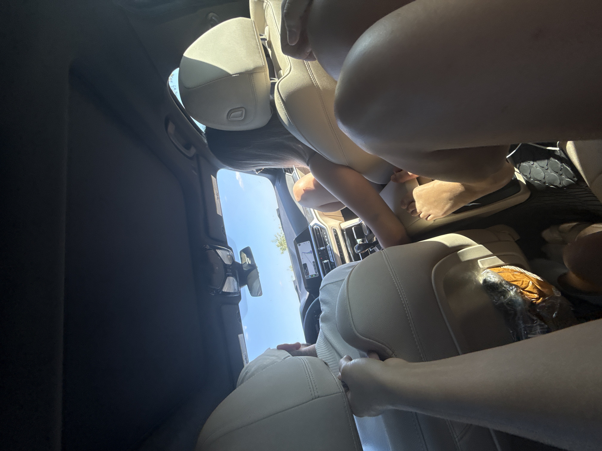 The image depicts the interior of a car from a rear-facing perspective. Two individuals are visible, both seated in the back. One person, to the left, is wearing a white sweater and is partially obscured by the headrest. The other person, to the right, is wearing a light-colored top and is looking forward. Their legs are prominently in the foreground, suggesting a close-up shot from within the car. Sunlight streams in from the side and front windows, illuminating the interior. The dashboard, steering wheel, and navigation screen of the car are visible. A bag of food items is also visible in a seat pocket. The image was taken in Fichitești, Romania.
