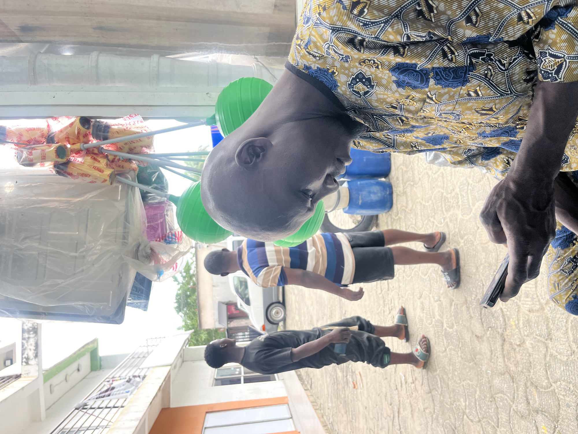 In Ado, Nigeria, on a paved outdoor area in front of a building with an orange and white facade, a bald African man in a yellow and blue patterned shirt is seated and looking down at his smartphone, which he holds in his left hand. In the background, two other African men are standing. One, wearing a black t-shirt and dark pants, is facing away from the camera. The other, in a blue and white striped shirt and dark shorts, is also facing away. To the left of the men is a white car. Above and behind the standing men, various items are hanging, including large green spheres and what appear to be cleaning supplies in yellow packaging. The scene suggests a commercial or residential area within the city.