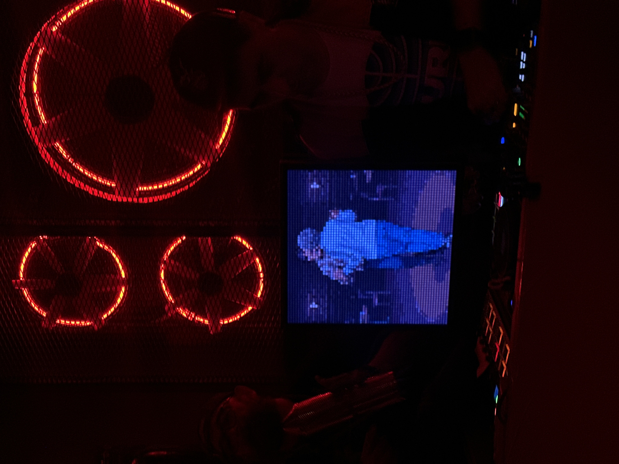 The image depicts the inside of a gaming computer case, illuminated with red LED lights. A large fan with a red LED ring is located at the top of the case. Below it are two smaller fans, also with red LED rings. A small monitor within the case displays a pixelated image of a person wearing blue clothing. A person's face is partially visible at the top of the frame, wearing a tank top with the lettering "JR." A keyboard with orange and red backlighting is positioned on the right side of the computer case. The environment is dark, with a reddish hue. The location is not identifiable based on the image alone.