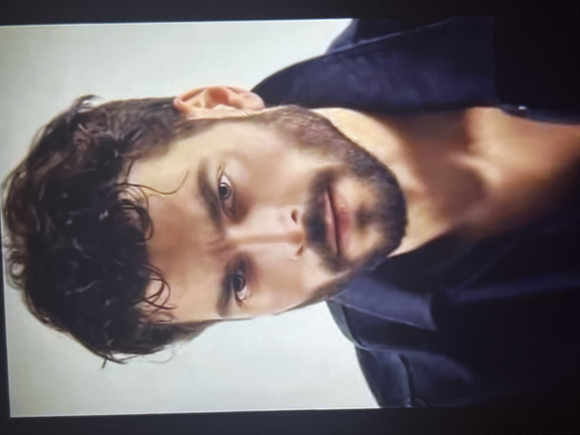 A medium shot captures a man with dark, curly hair and a beard. He's wearing a dark, buttoned shirt or jacket. The background appears to be a plain white wall. The image seems to be taken indoors, but the exact location in an unknown city is not discernible from the visual information provided.