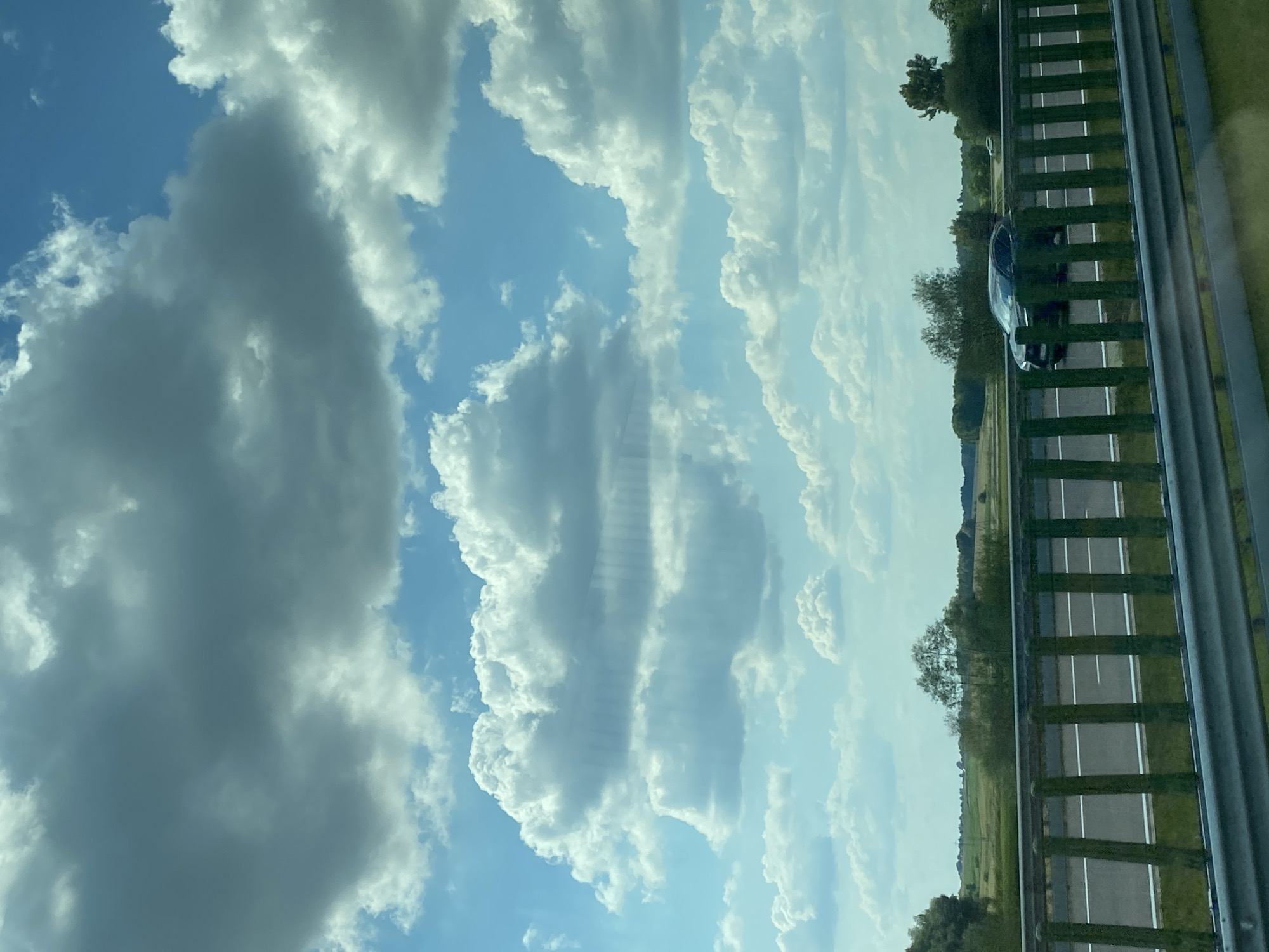 The image captures a view from a vehicle traveling on a highway in Dobkowice, Poland, under a partly cloudy sky. The foreground features a green metal guardrail, with the highway stretching ahead. A dark-colored sedan is visible on the highway. In the background, a flat, green landscape with scattered trees and fields extends to the horizon. The sky is a mix of bright blue and large, white, and gray clouds.