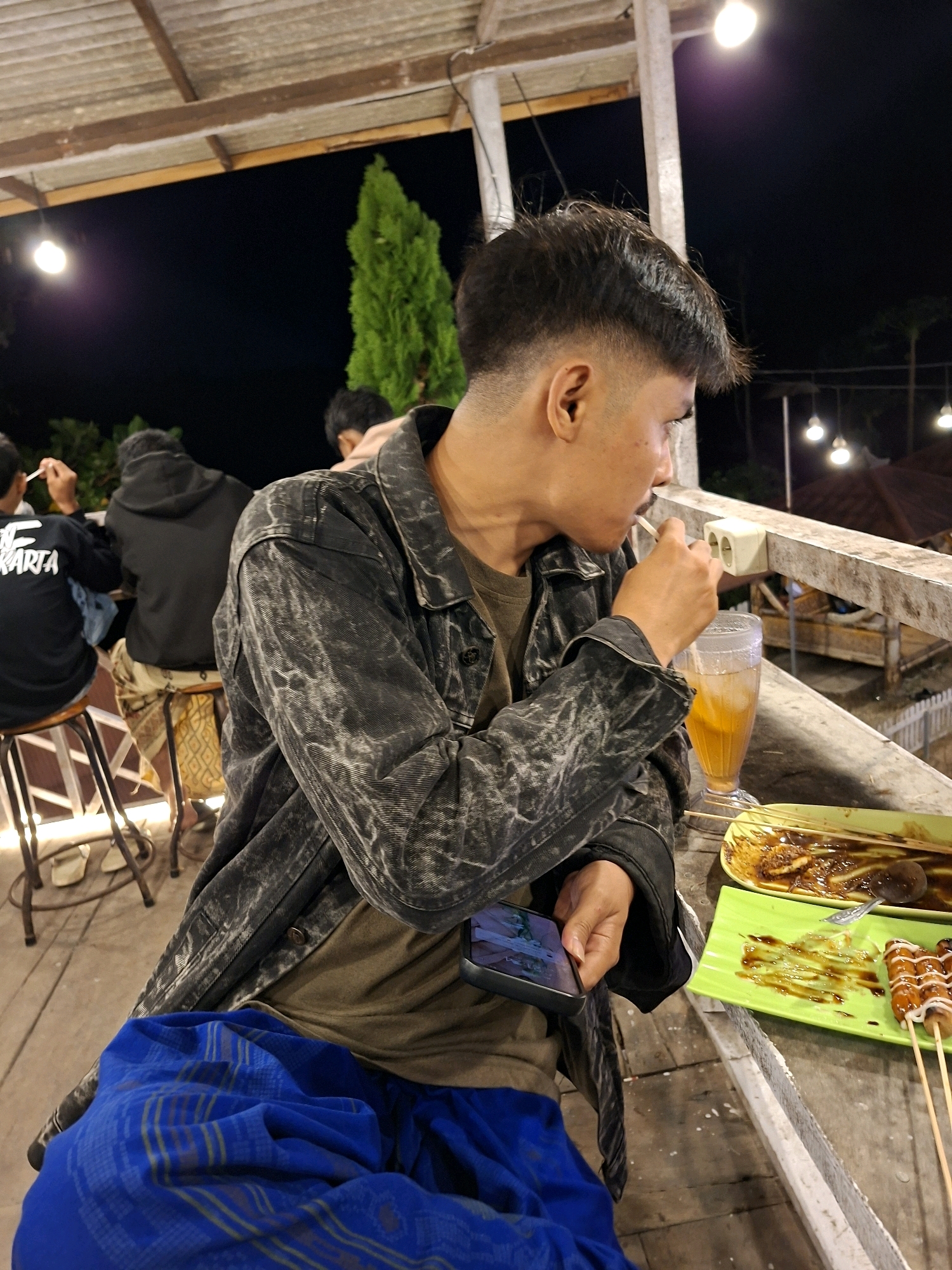 In Lantek Barat, Indonesia, at night, a young man with short, styled dark hair sits at an outdoor table, looking to his right. He wears a dark, distressed denim jacket over a t-shirt, and a vibrant blue sarong covers his lap. In his right hand, he holds a straw to a tall glass of iced beverage, while his left hand rests on his phone, which displays an image. The table surface, a weathered wooden deck, has a green plate with skewered sausages drizzled with sauce and mayonnaise, and another plate with remnants of food and sauce. Behind the man, other individuals are visible, one in a black hoodie and another in a black shirt with white lettering. The environment is dimly lit by overhead lights and features a rustic wooden roof structure.