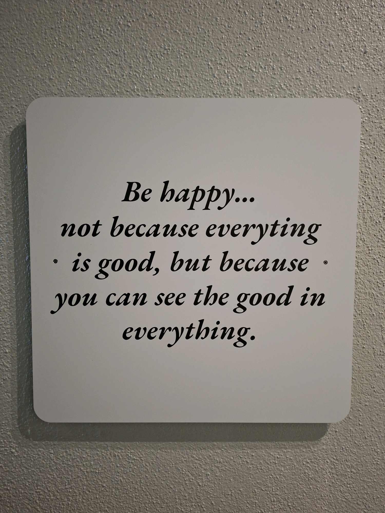 A white rectangular sign with rounded corners is mounted on a textured, light gray wall. The sign contains black text in a script font. The text reads: "Be happy... not because everything is good, but because you can see the good in everything." The sign casts a shadow on the wall below it. There are no people or animals visible in the image. The location is Olgiate Olona, Italy.
