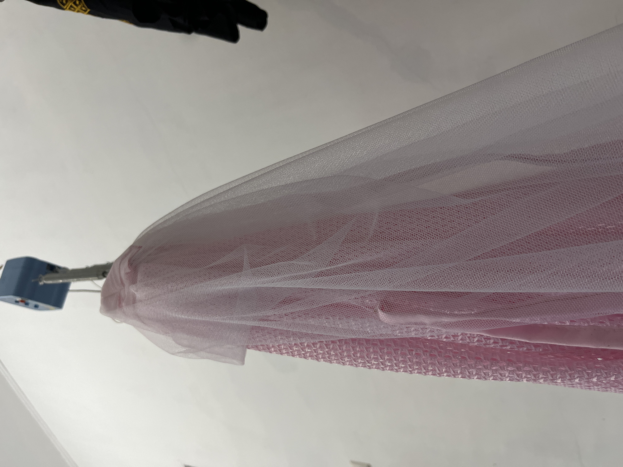 The image displays an indoor scene featuring textile materials and a mounting device. The shot is oriented such that a blue rectangular device is positioned on the left side of the frame, affixed to a light-colored wall or ceiling. Metal rods extend from this device to the right, supporting a large quantity of draped fabric. The fabric consists of multiple layers: an outer layer of light pink to white sheer mesh, possibly tulle or netting, which is draped across the middle and right portions of the image. Beneath this, layers of solid pink fabric and a distinctively textured, patterned pink material (resembling bubble wrap or a honeycomb pattern) are visible. In the top left corner of the frame, a dark object, potentially a glove or piece of apparel, is partially visible, featuring a yellow, somewhat abstract pattern that could be a logo or text, though it is indistinct. The background is a plain, light gray or white surface. The scene is static, suggesting a display, storage setup, or manufacturing context for textiles. Lighting appears bright and even, consistent with an indoor environment in Lhokseumawe, Indonesia.