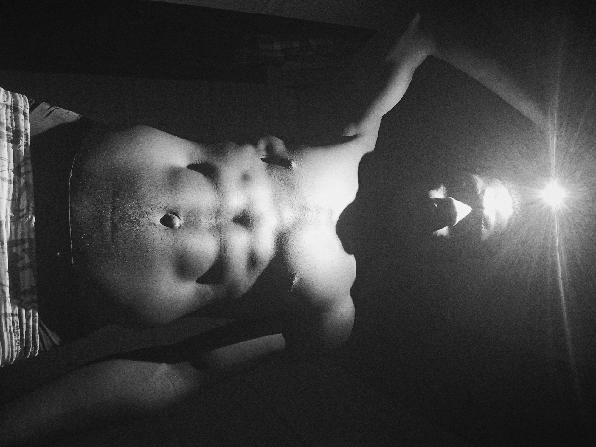 A black and white, high-contrast, low-angle photograph features a partially nude male torso lying on what appears to be a bed. The subject is positioned with his head near the top right corner of the frame and his feet towards the bottom left. His torso is well-defined with visible abdominal muscles. A bright light source shines directly onto his face from the upper right, creating a lens flare. The background is mostly dark and indistinct. Fabric, possibly bedding, is visible on the left side of the frame. The location, Nkpor, Nigeria, is not evident in the image.