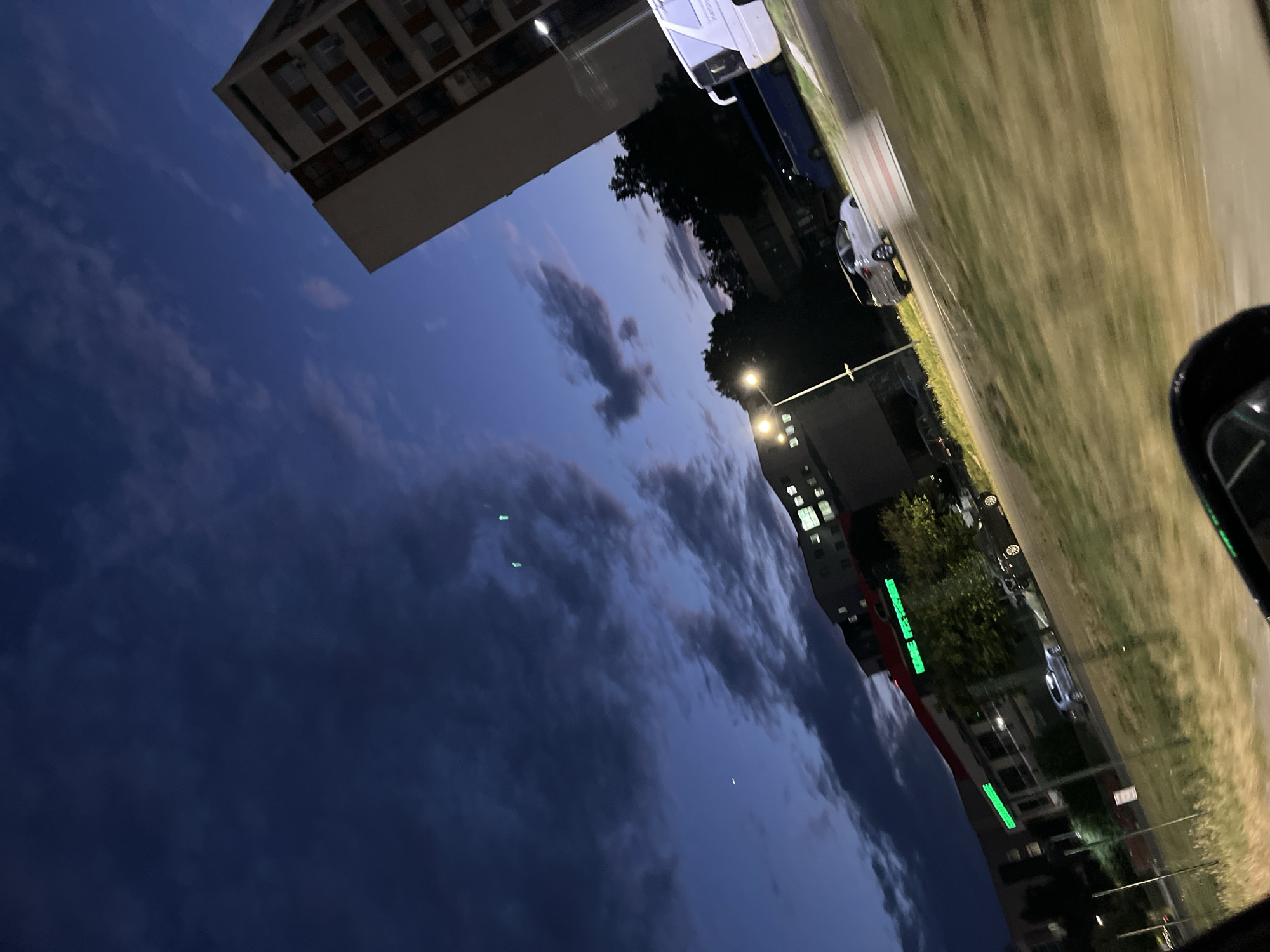 The image depicts a street scene at dusk or dawn, with a dark, cloudy sky. The perspective is from inside a vehicle, looking out through the window, as evidenced by the reflections and the car's side mirror in the lower right corner. Buildings are visible on the left and in the background, with lights on in some windows. The street is lined with grass and trees. Several cars are parked or driving on the street, including a silver sedan and a white van. There is a bright green illuminated sign on one of the buildings, which appears to have text on it, though it is partially obscured and illegible. The overall atmosphere is calm and quiet. The orientation of the image suggests it may have been taken while the vehicle was moving.