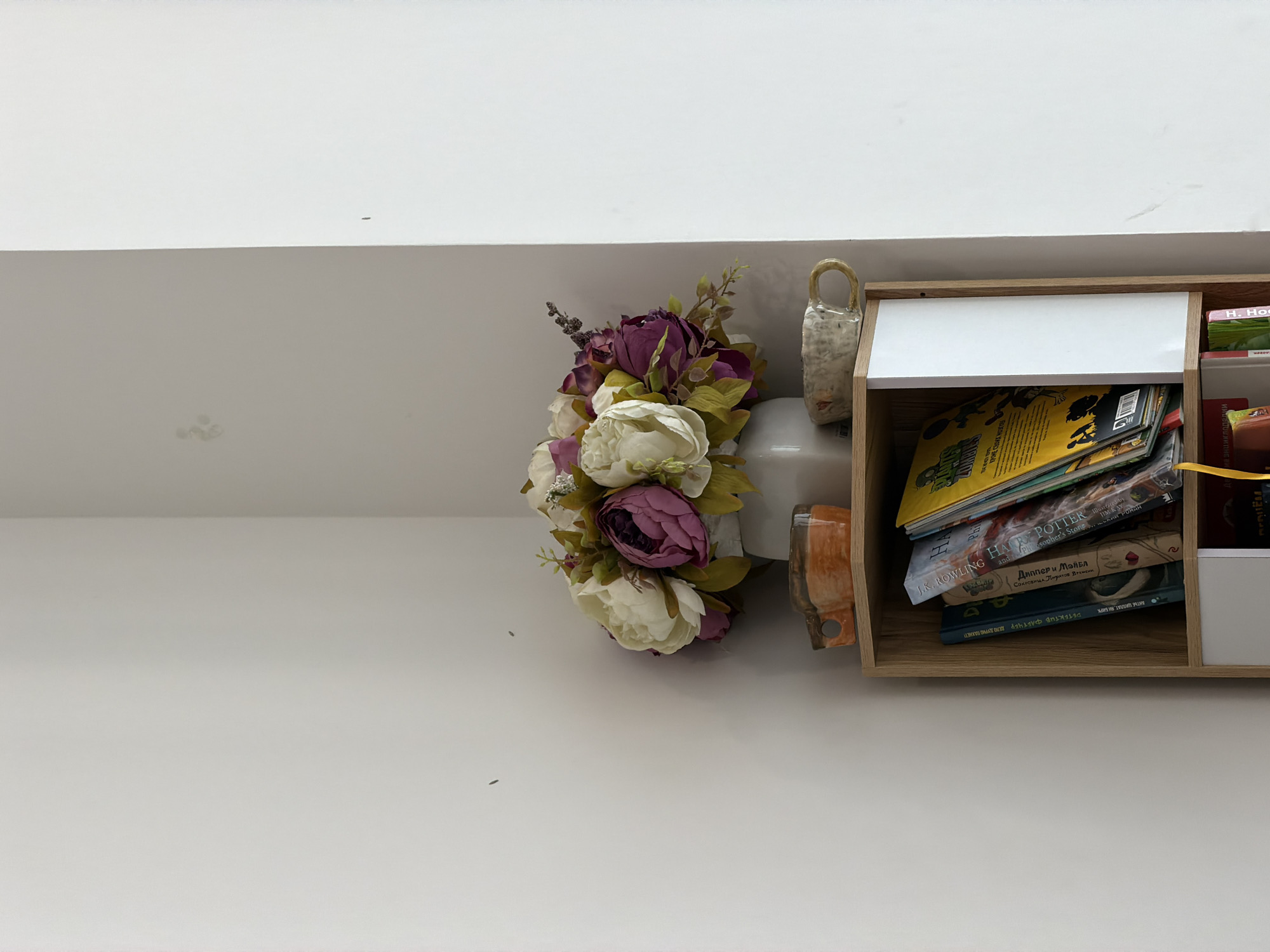 This image captures a detail of an indoor setting, focusing on a wall-mounted shelf unit, decorative flowers, and a section of a wall and ceiling. The main subject is a light-colored wooden shelf, which contains several books and comic books stacked horizontally.
Visible text on the books includes "JK ROWLING HARRY POTTER and the Philosopher's Stone," suggesting a copy of the popular fantasy novel. Other titles in Russian include "Диппер и Мэйбл: СОКРОВИЩА Пиратов Времени" (Dipper and Mabel: Treasures of the Pirates of Time), and "ДЕТЕКТИВ ФАРЕТЧЕР ДЕЛО ДУРНО ПАХНЕТ" (Detective Fartcher: The Case Smells Bad) by "АНТЬЕ ЦИЛЛАТ, ЯН БИРК" (Antje Damm, Jan Birck). Partial texts like "H. Ho" and "на энциклопедии" are also present.
Adjacent to the shelf is a decorative bouquet of artificial flowers, primarily white, cream, and purple peonies, arranged in a vase. A small, textured, off-white ceramic or clay object with a handle is placed near the flowers, leaning against the shelf.
The background consists of a clean white surface, likely a ceiling or an upper portion of a wall, above a lighter, possibly light gray, wall. Both surfaces show minor scuffs, dirt marks, and small dark specks. The overall scene depicts a small, decorated storage area within what appears to be a home in Bac Nha Trang Ward, Vietnam. The image itself is presented in a sideways orientation.